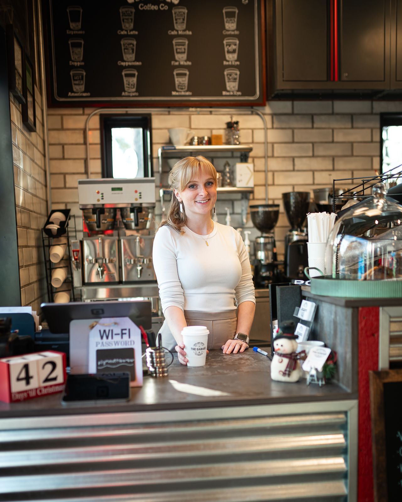 A barista stands behind the counter at a coffee shop with exposed brick walls and menu board displaying drink options.