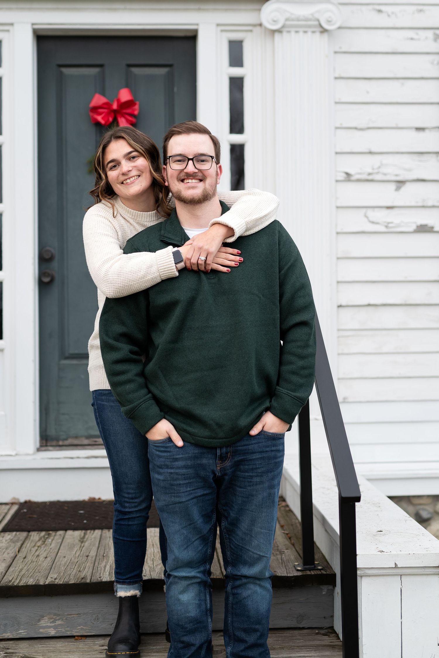 Couple in casual sweaters standing on white porch with red bow decoration.