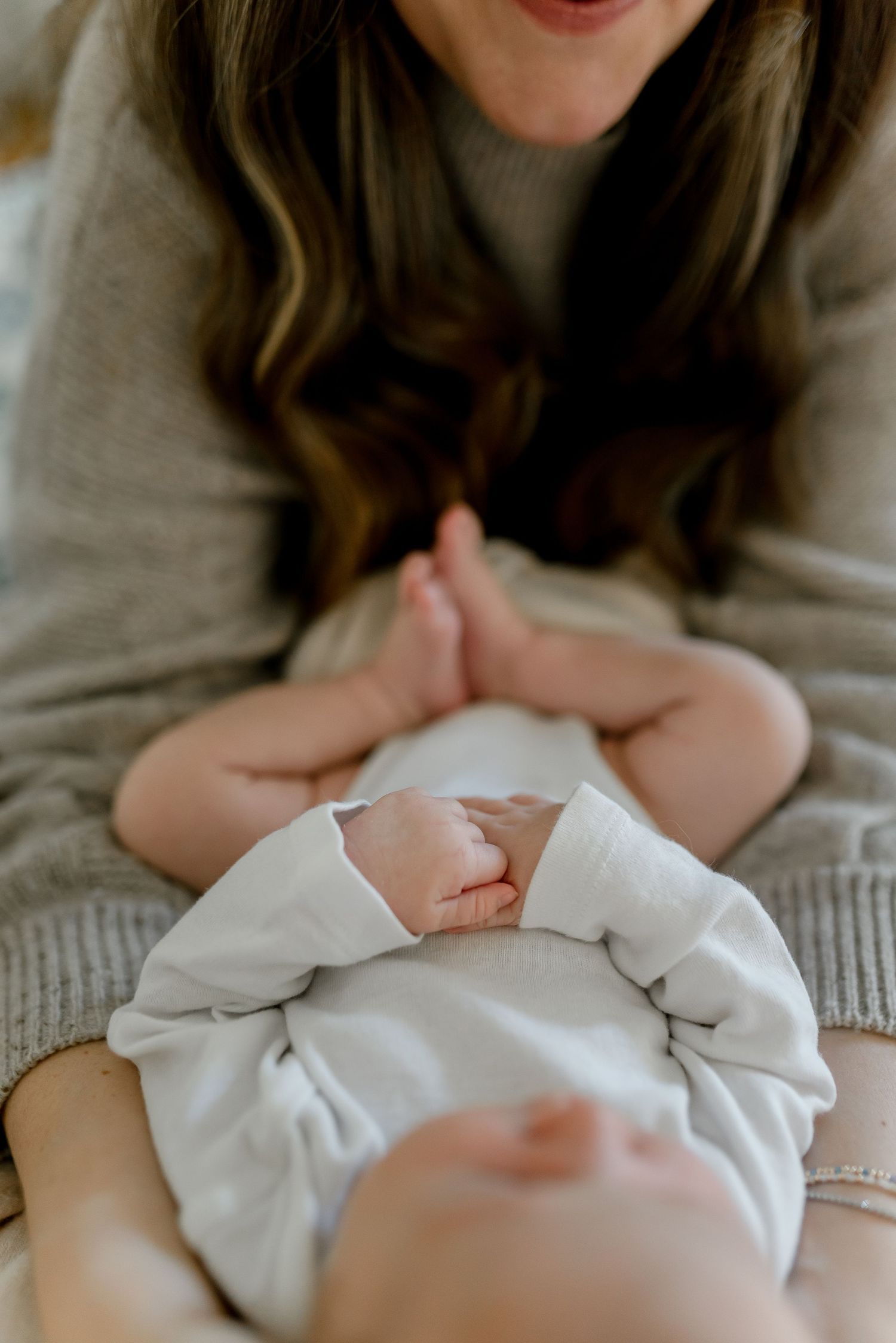 Mother tenderly holding newborn baby's tiny hands while leaning over infant in white onesie.