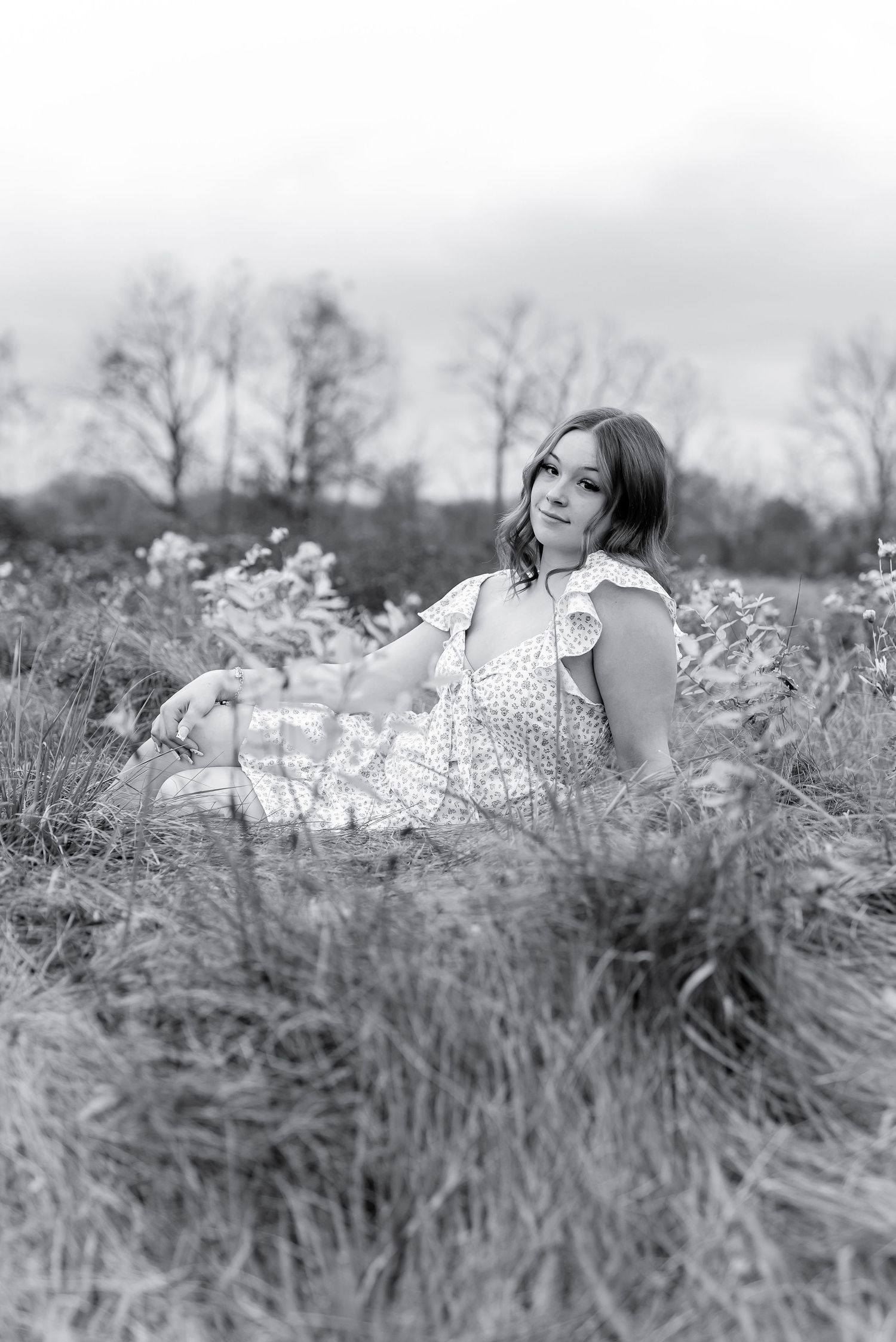 Black and white photo of young woman in floral dress sitting in tall grass field.