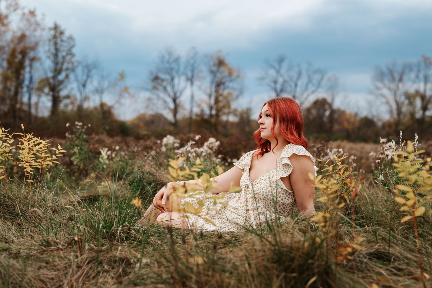 Red-haired woman in floral dress sitting peacefully in autumn meadow with wildflowers and cloudy sky.