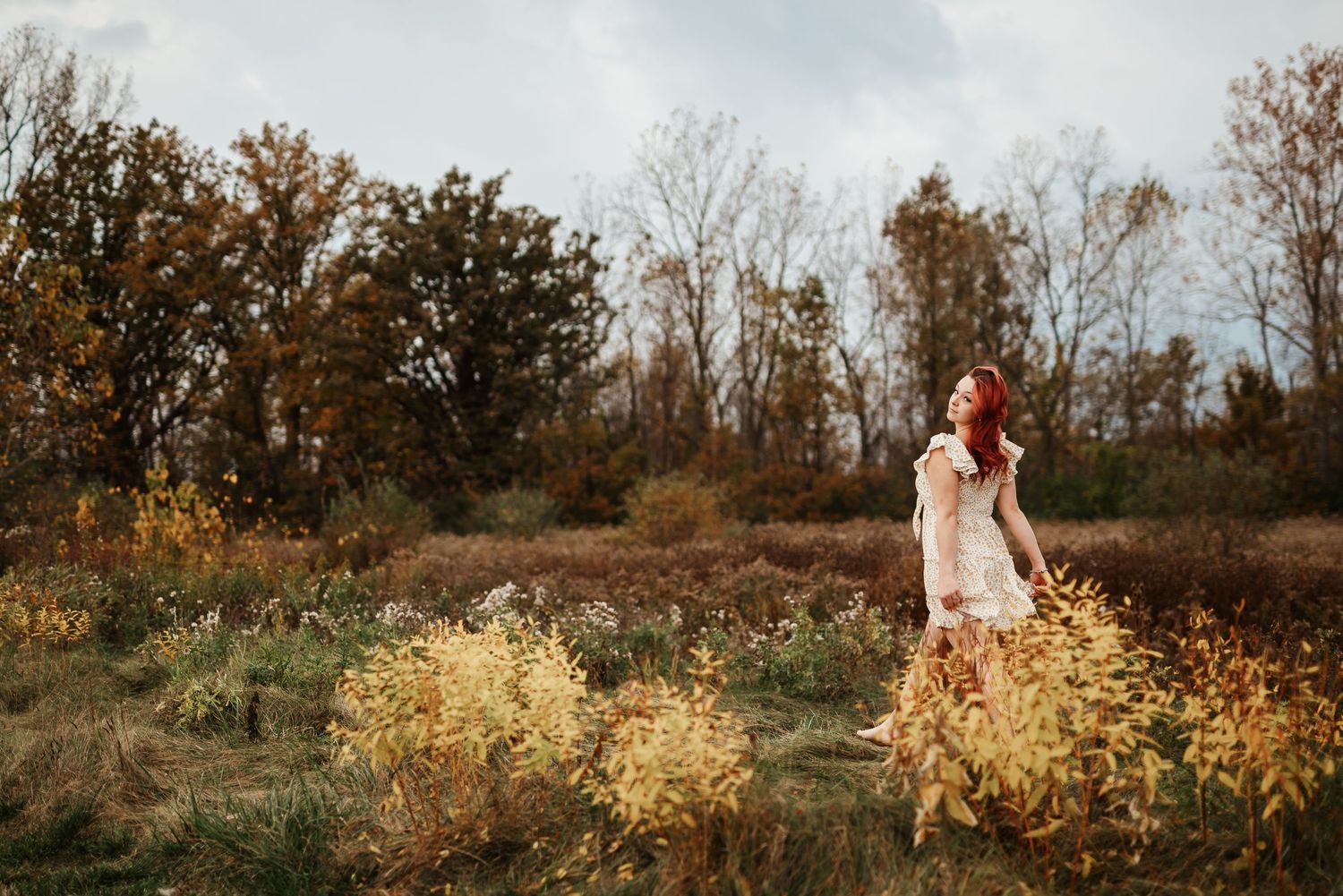 A woman in a floral dress stands among autumn wildflowers in a golden field with colorful fall trees behind her.
