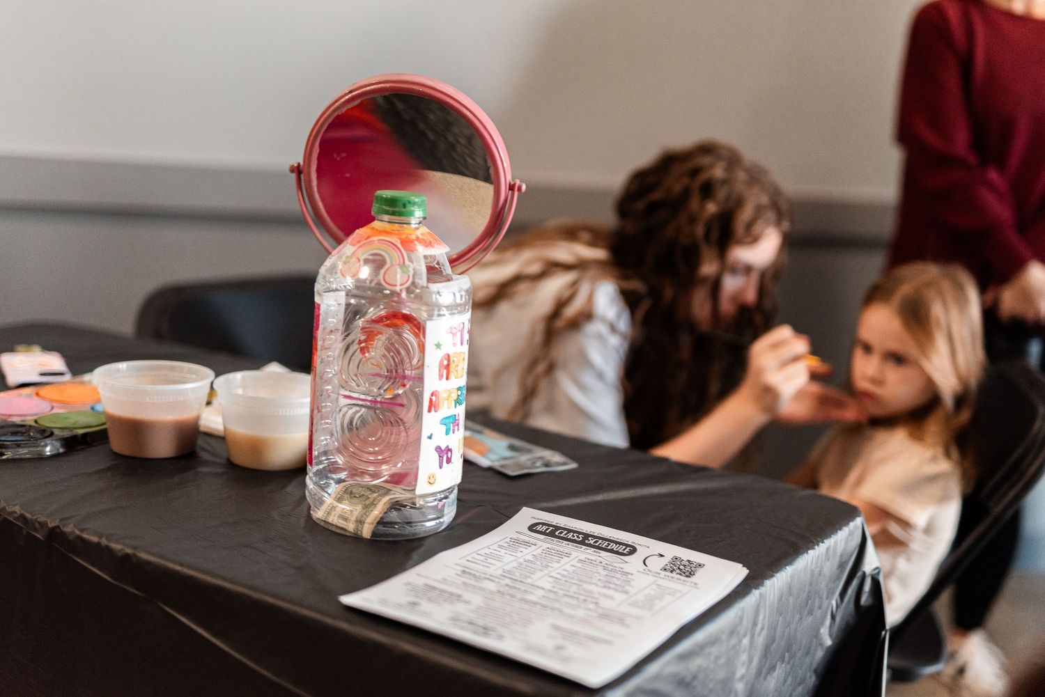 Child getting face paint at a Halloween activity table with craft supplies and water bottle.