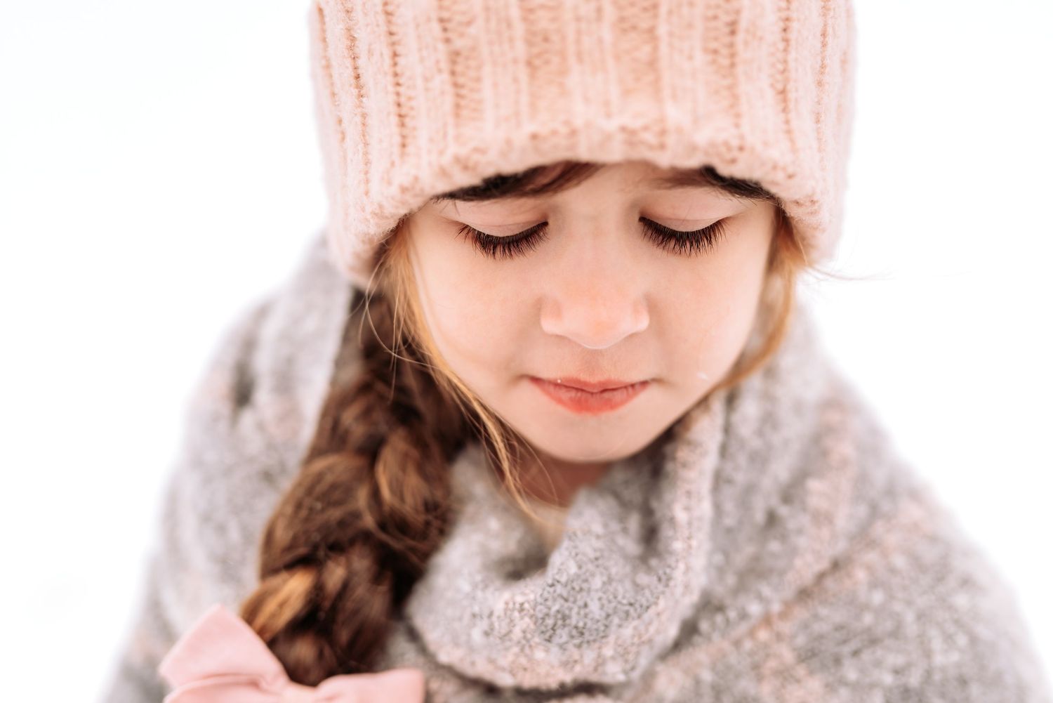 A child in a cozy knit hat and winter sweater smiles while looking down in a soft, dreamy portrait.