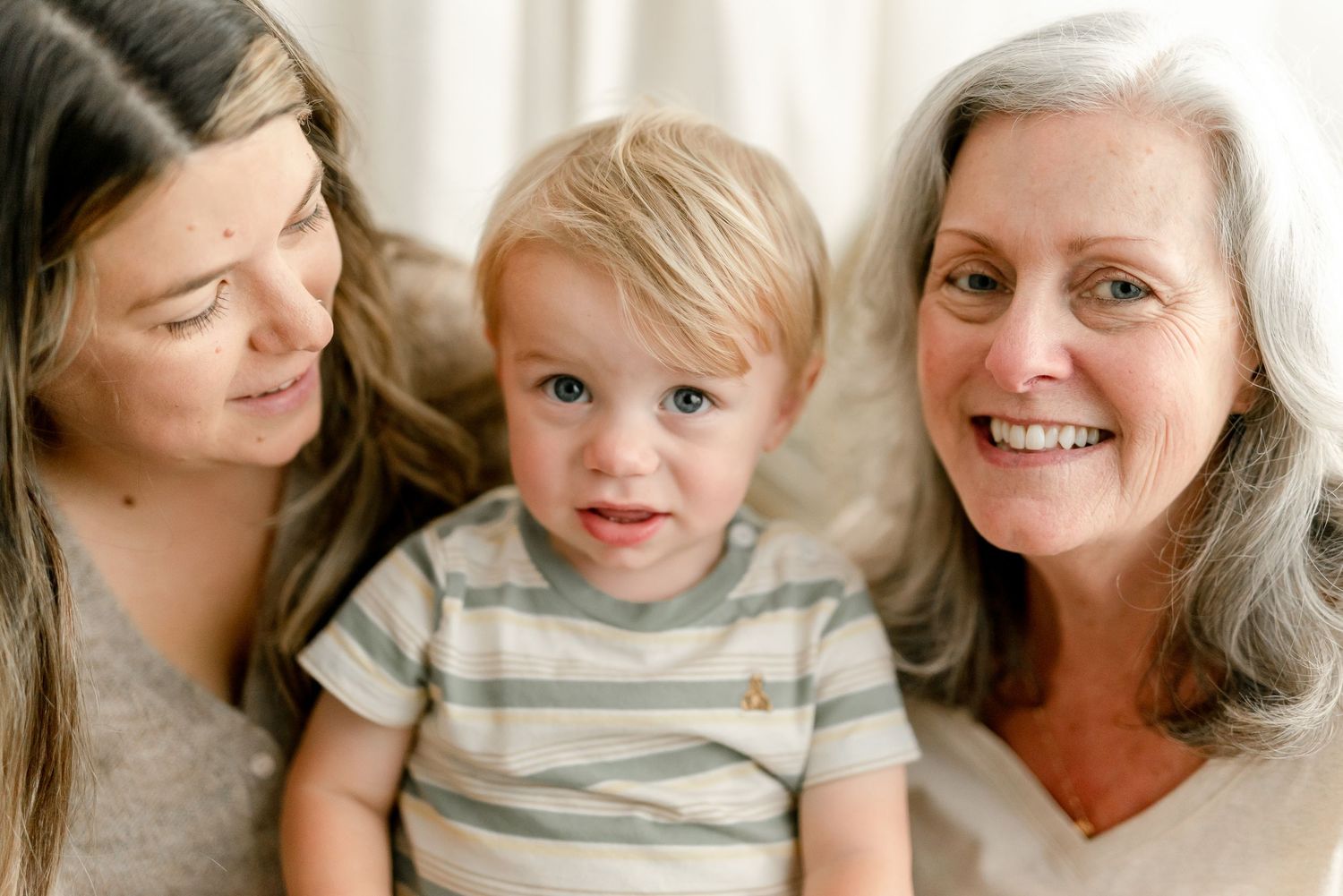 Smiling young woman and gray-haired older woman sit close together holding a happy toddler in a striped shirt.