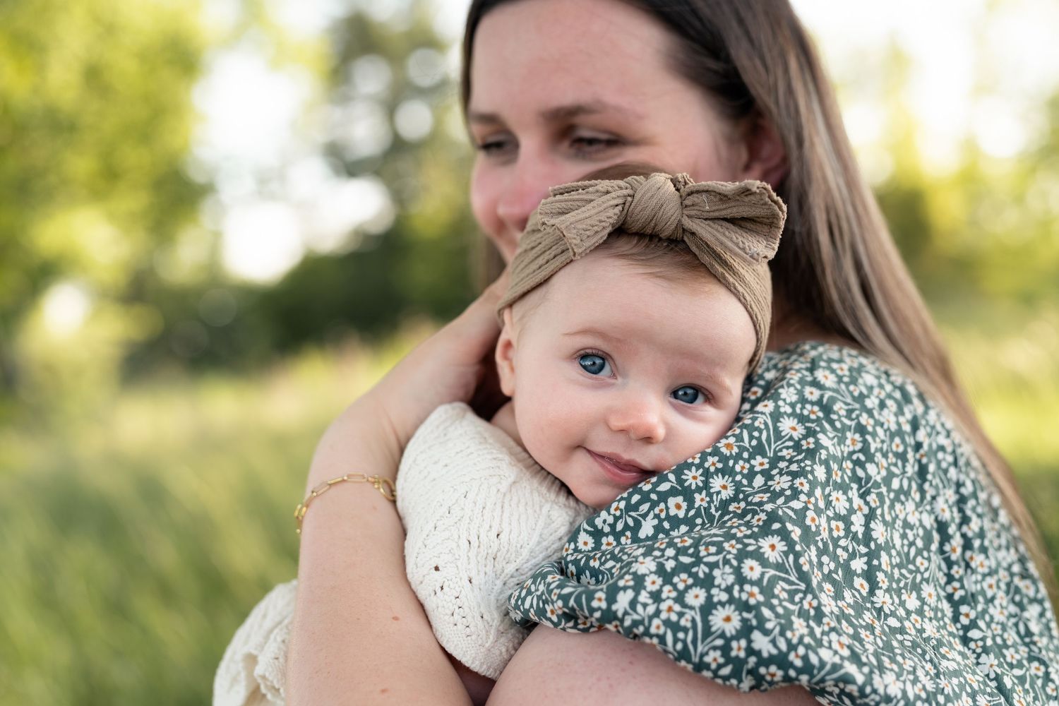 A mother tenderly holds her blue-eyed baby girl wearing a bow headband outdoors in a green field.