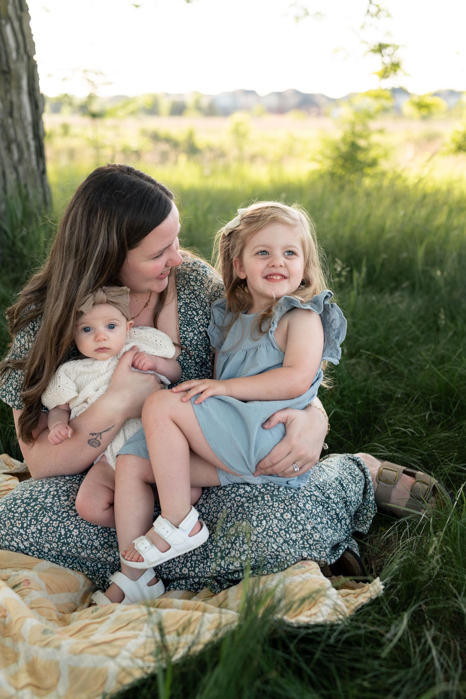 A mother sits outdoors in a floral dress holding her baby and toddler daughter in a lush green field at golden hour.