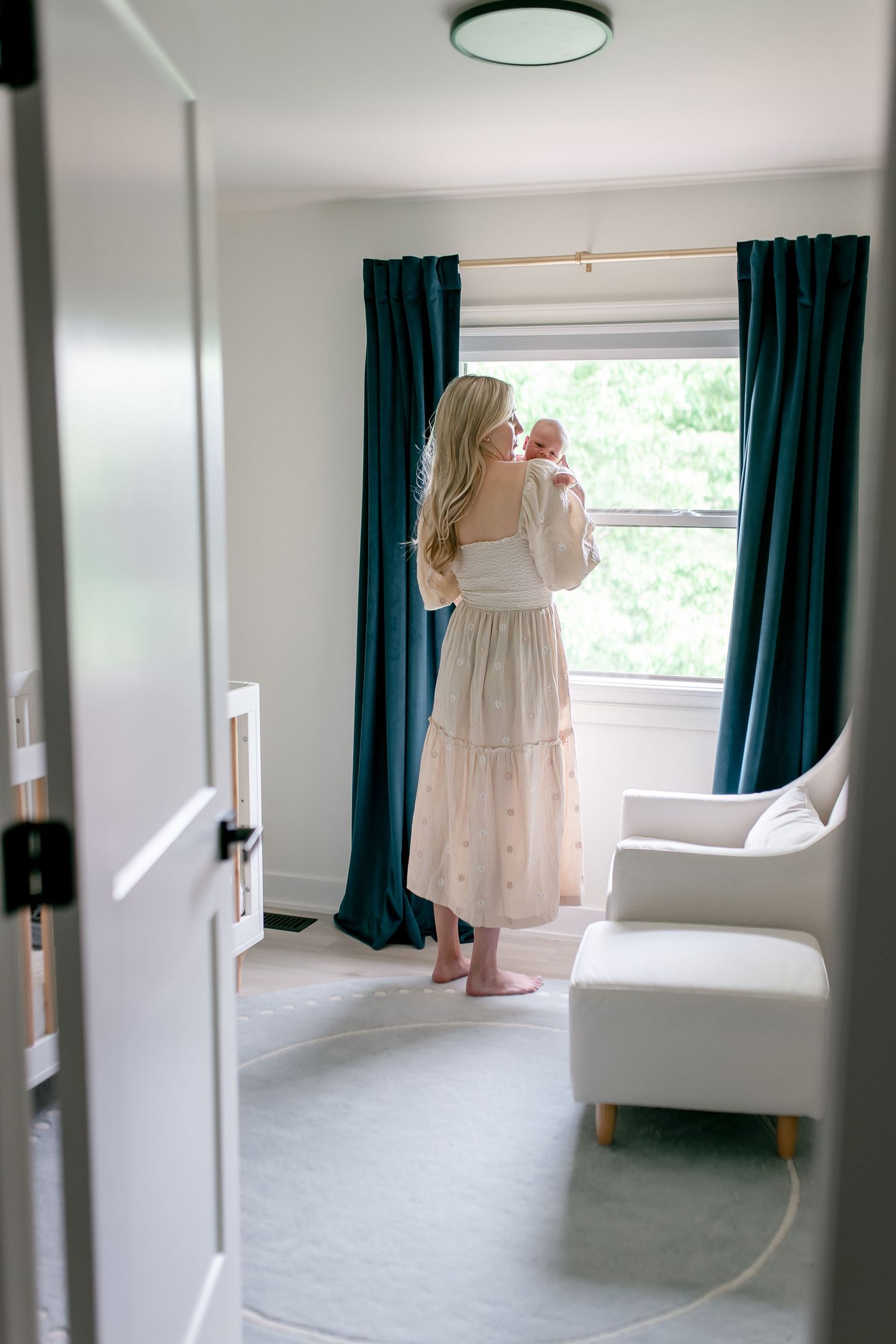 A person in a white tiered dress stands by a window with teal curtains in a bright minimalist room.