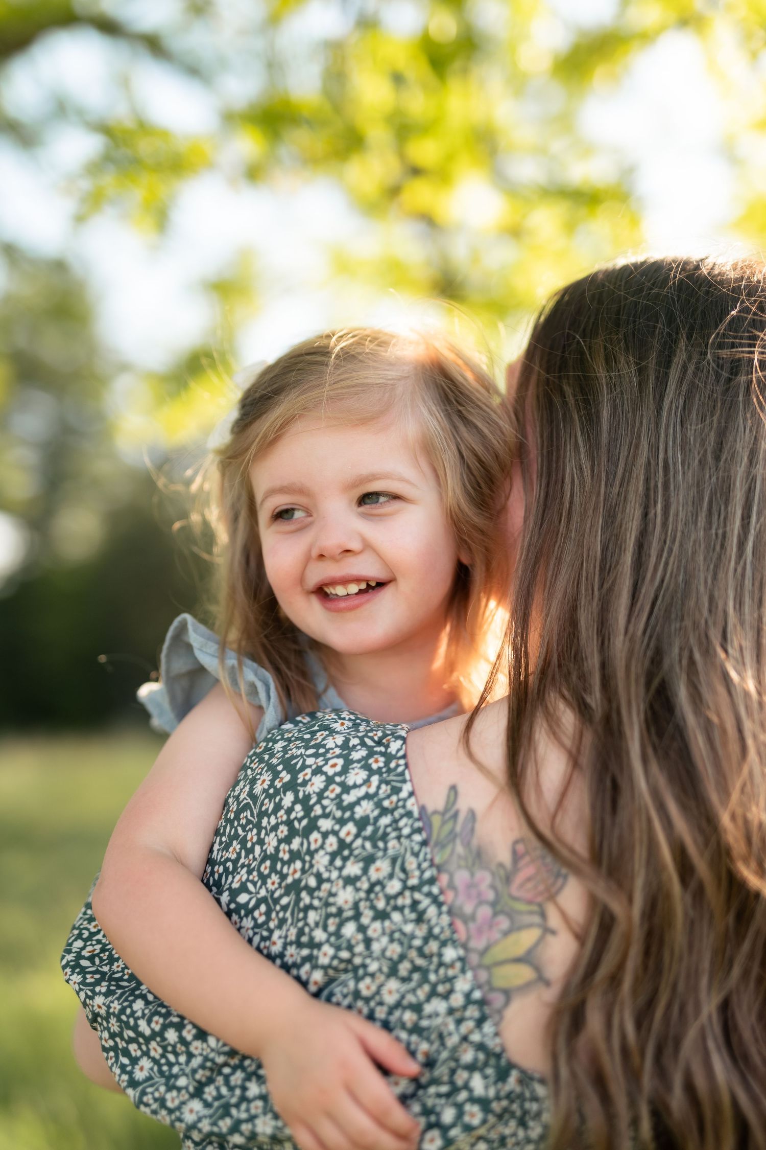 A mother with a floral tattoo kisses her smiling toddler girl in a sunny green park.