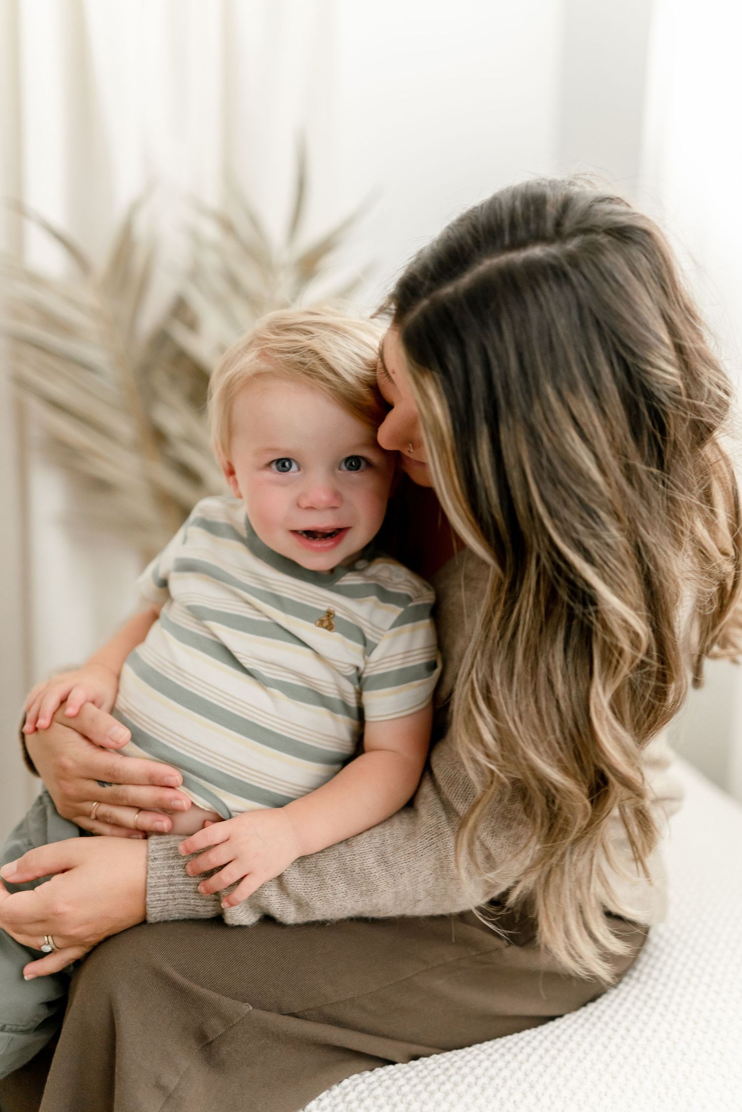 A woman with long wavy hair gently kisses a smiling blond toddler in a striped shirt while holding him in her lap near dried palm fronds.