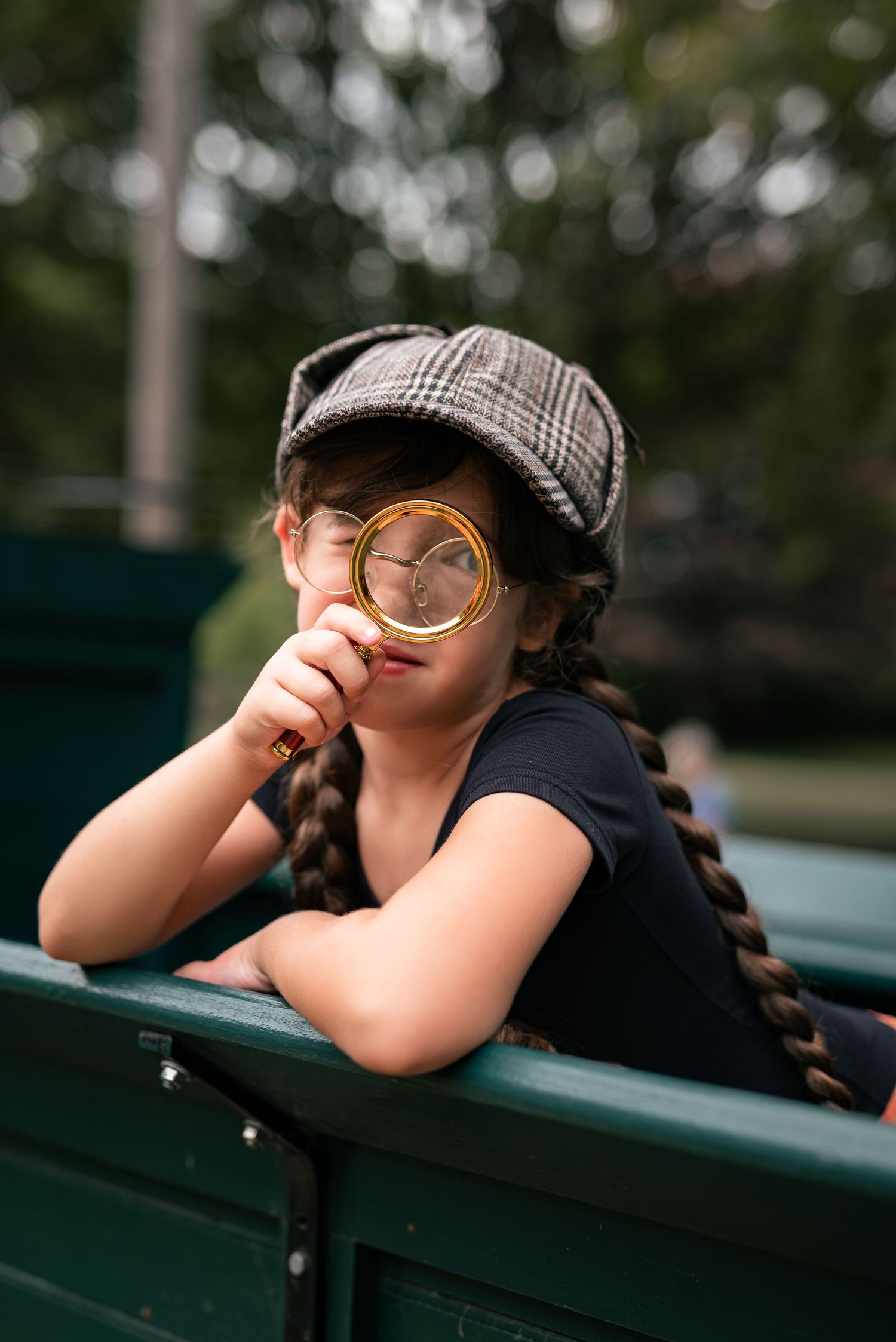 Young girl in plaid cap peers through magnifying glass while leaning on green bench outdoors.