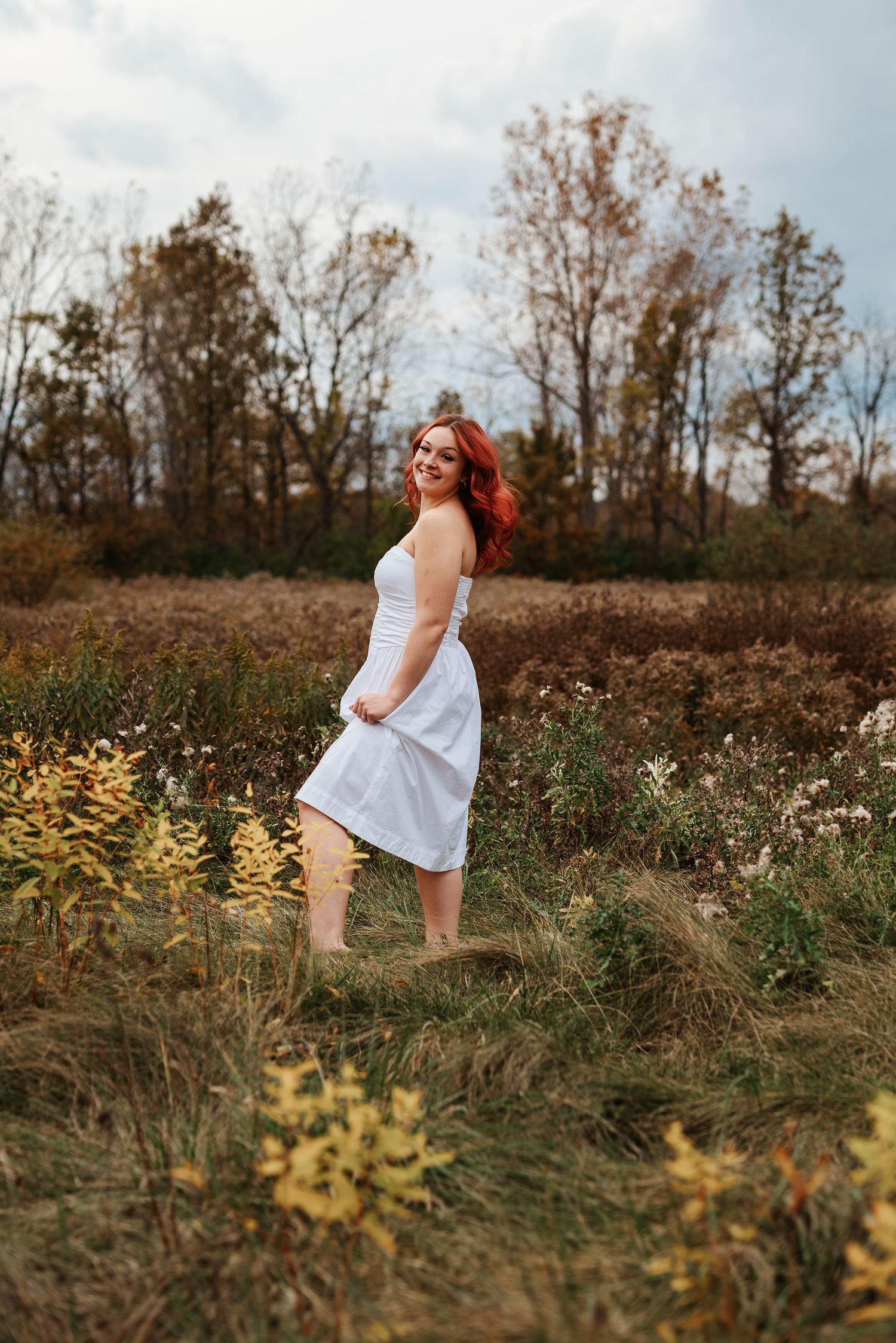 Red-haired woman in white dress twirling in autumn field with wildflowers and trees.