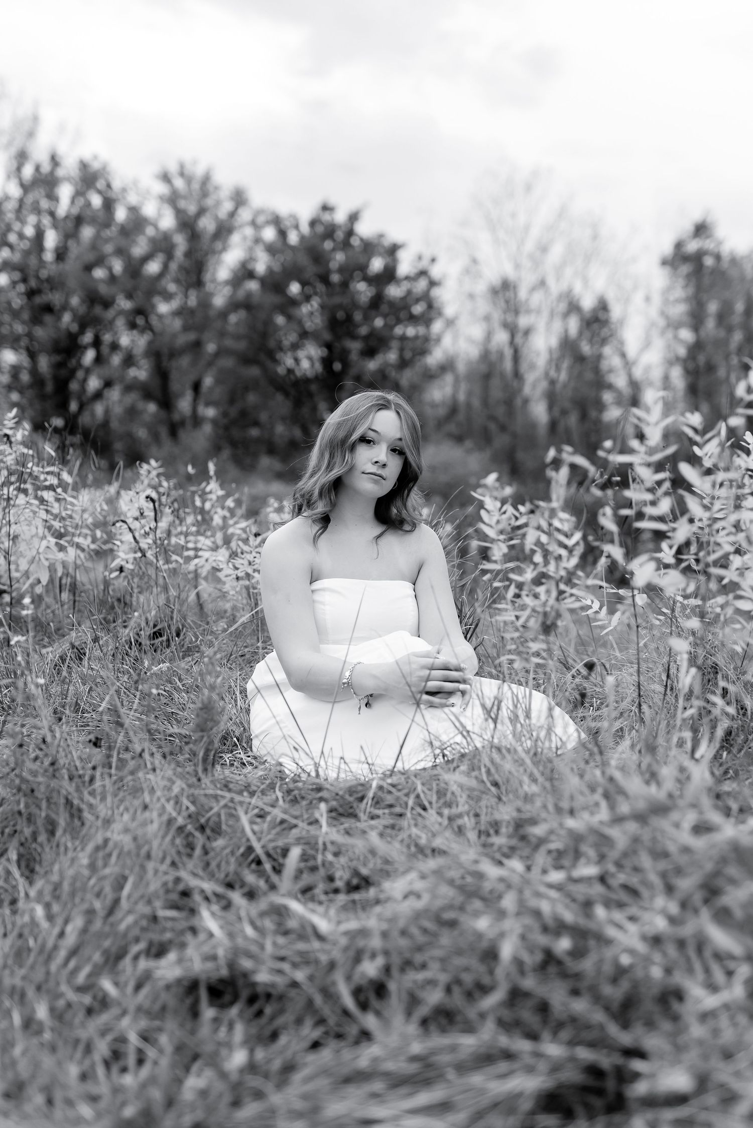 Black and white portrait of a woman in white dress sitting peacefully among tall grass and wildflowers outdoors.