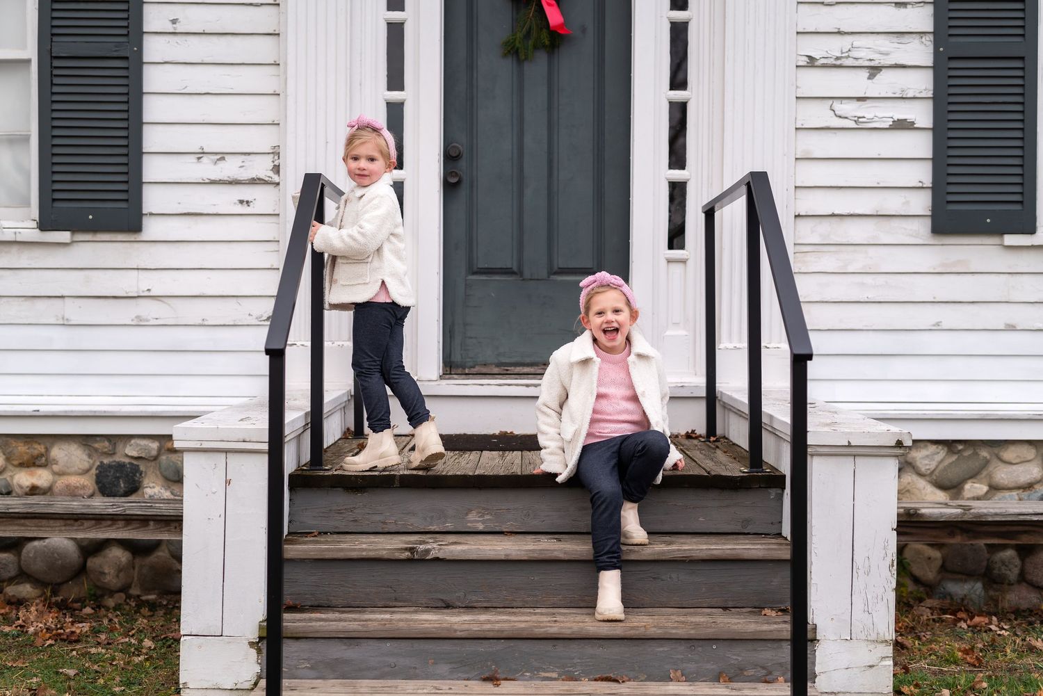 Two children in matching cream sweaters playing on wooden steps of a white house with a holiday wreath.