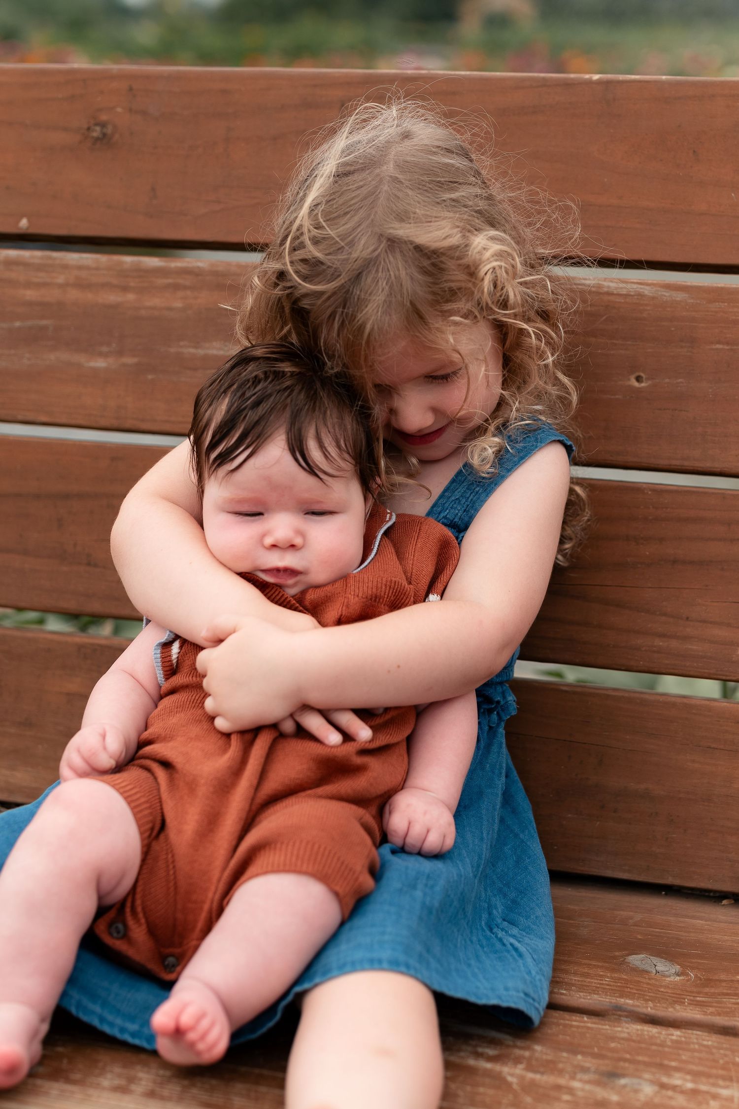 Young girl with curly hair hugging a baby sibling on a wooden bench outdoors.
