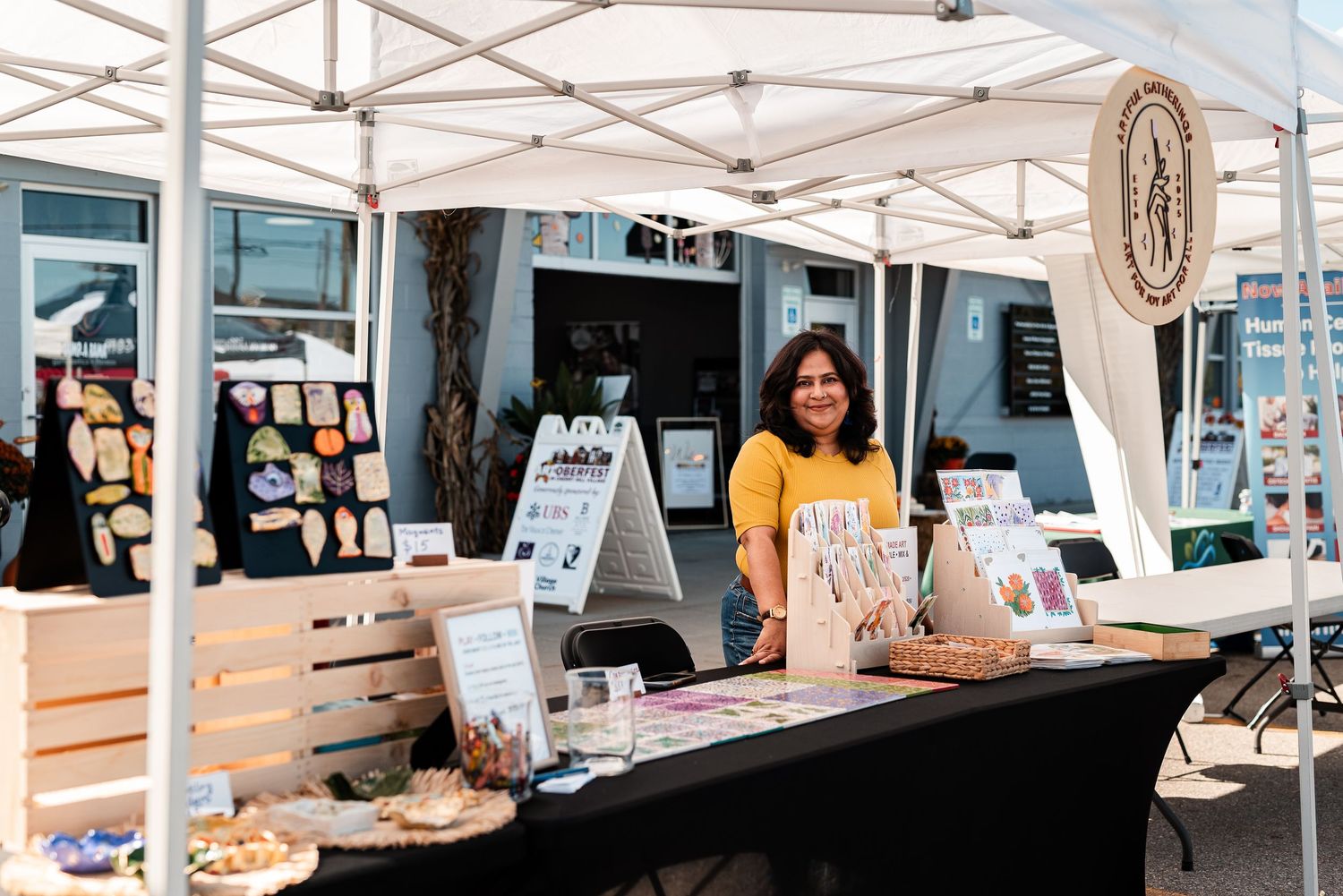 Woman in yellow shirt smiling at outdoor market booth with crafts and products displayed under white canopy tent.
