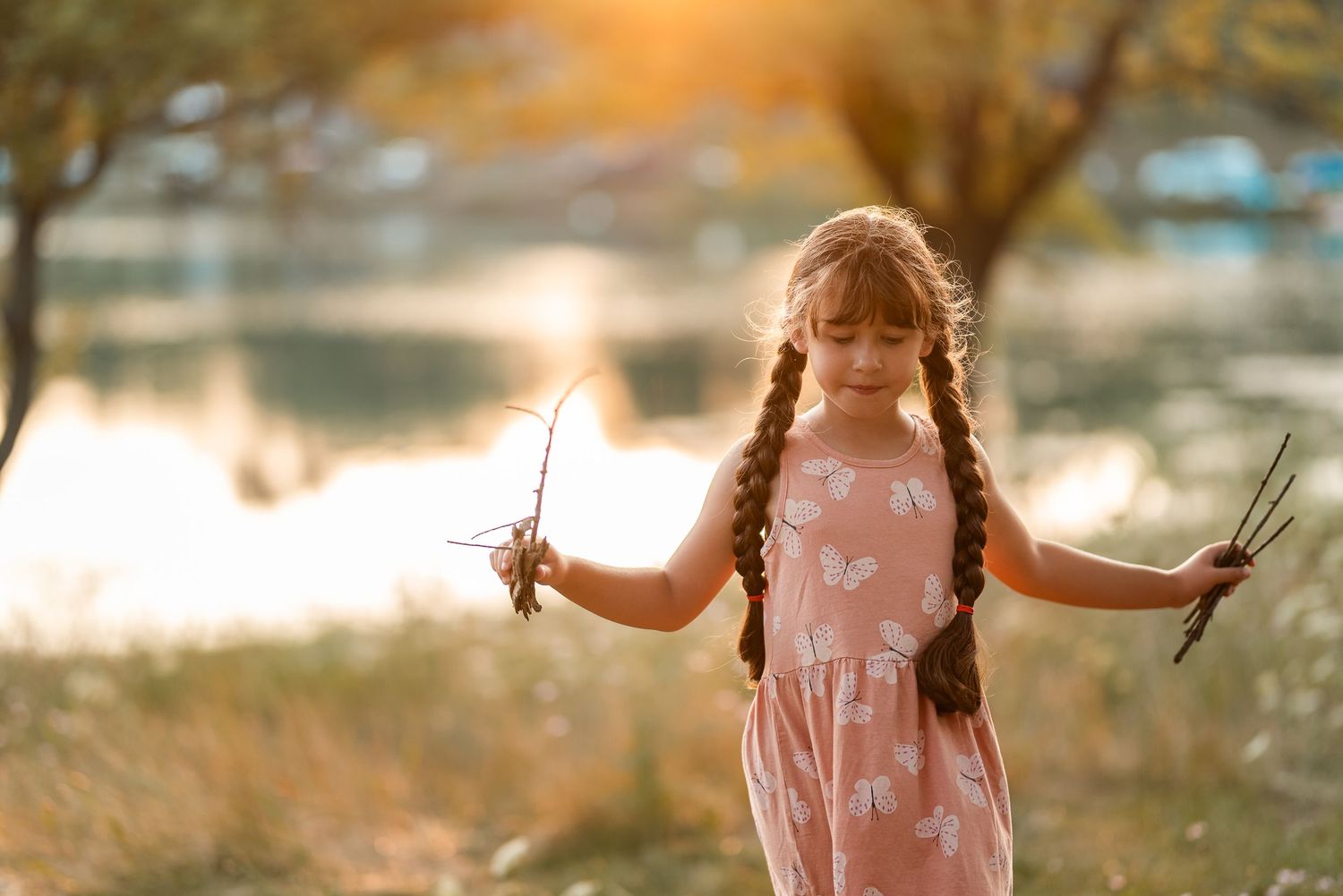 Young girl with braids in pink dress holds sticks outdoors near lake at golden sunset.