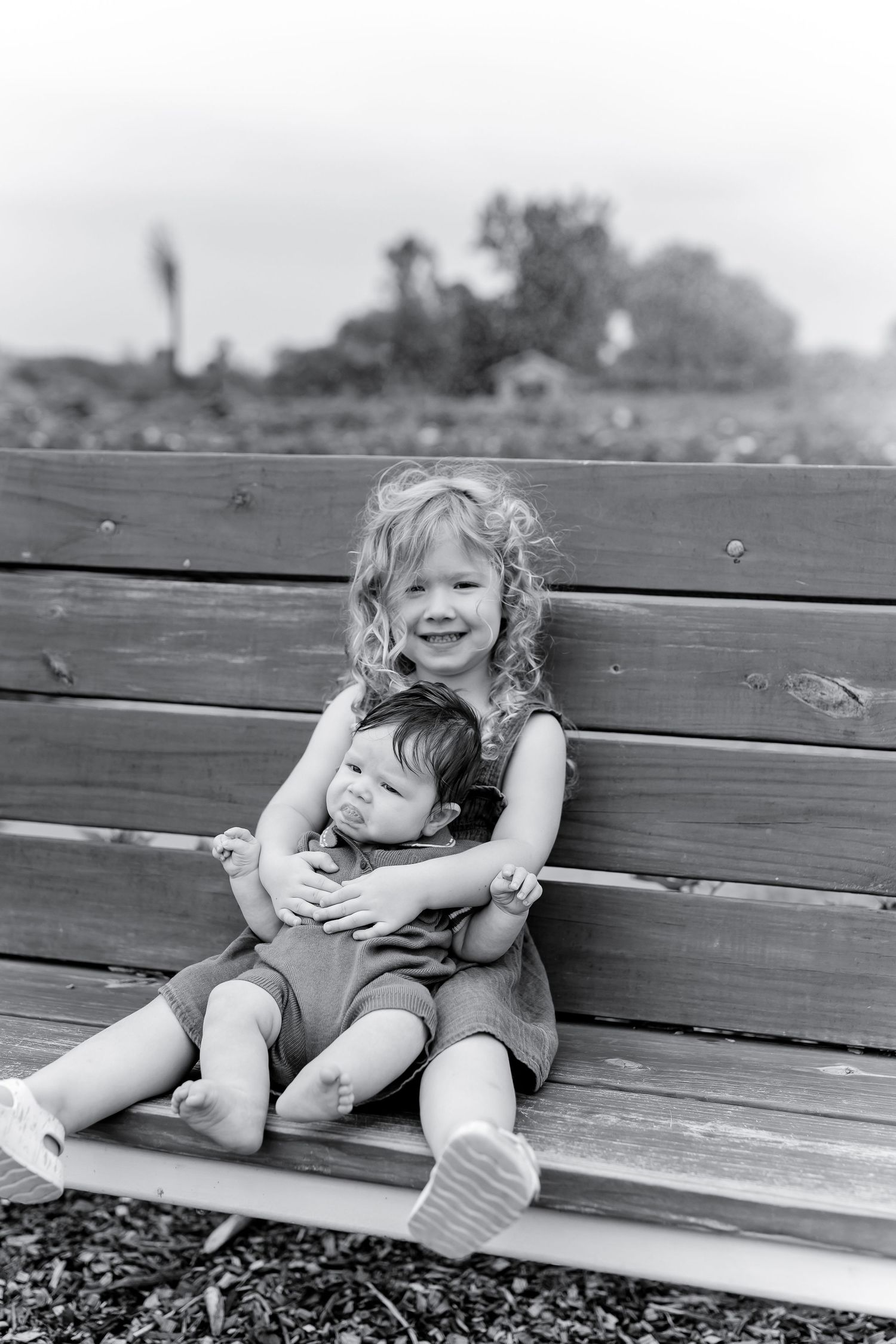 A smiling girl with curly hair hugs a toddler on a wooden bench outdoors in a black and white photo.