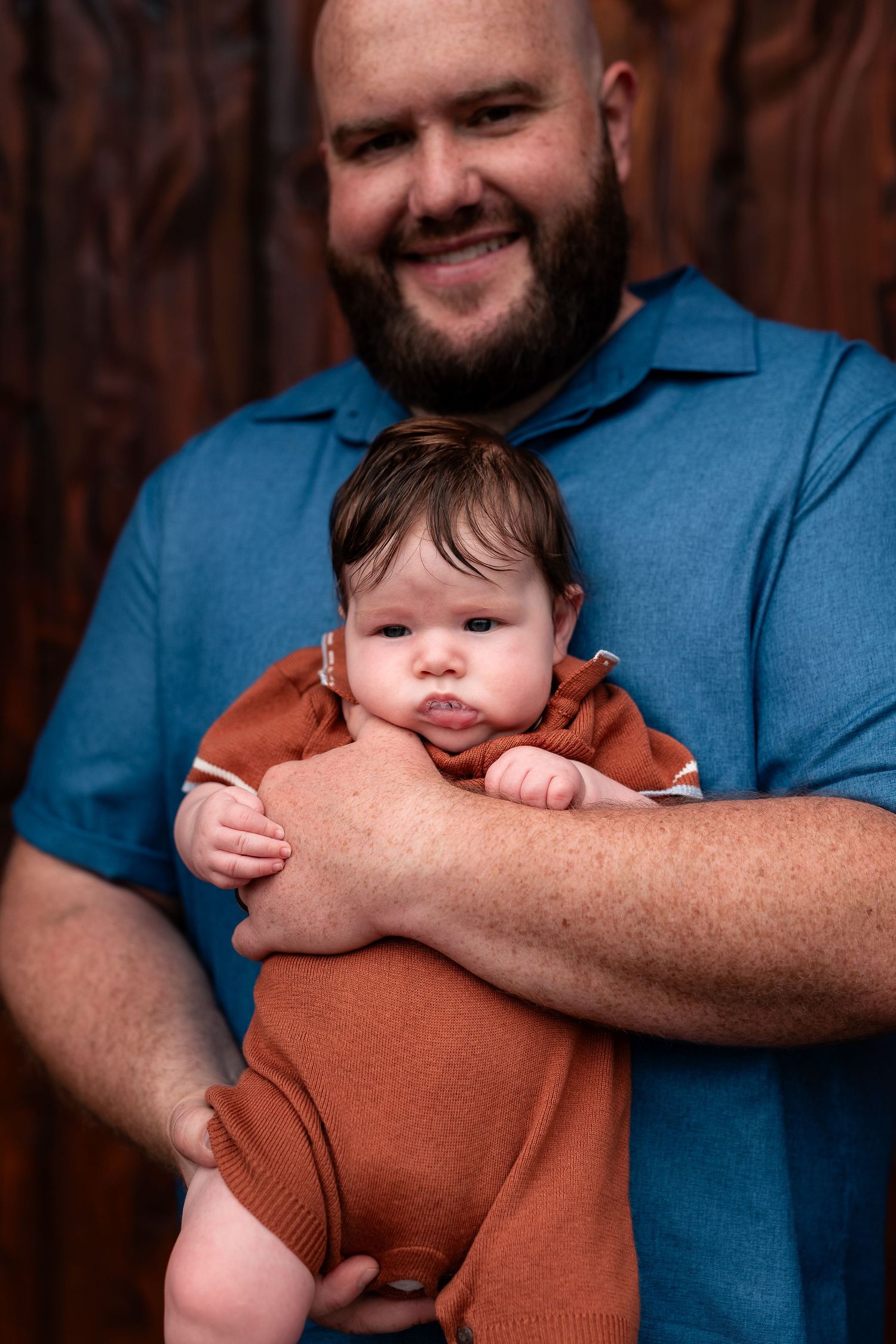 Smiling bearded man in blue polo shirt holding a newborn baby in a rust-colored romper against a wood backdrop.