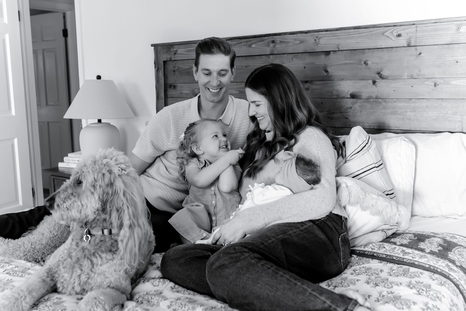 Black and white photo of parents smiling at newborn baby with toddler and fluffy dog on bed.
