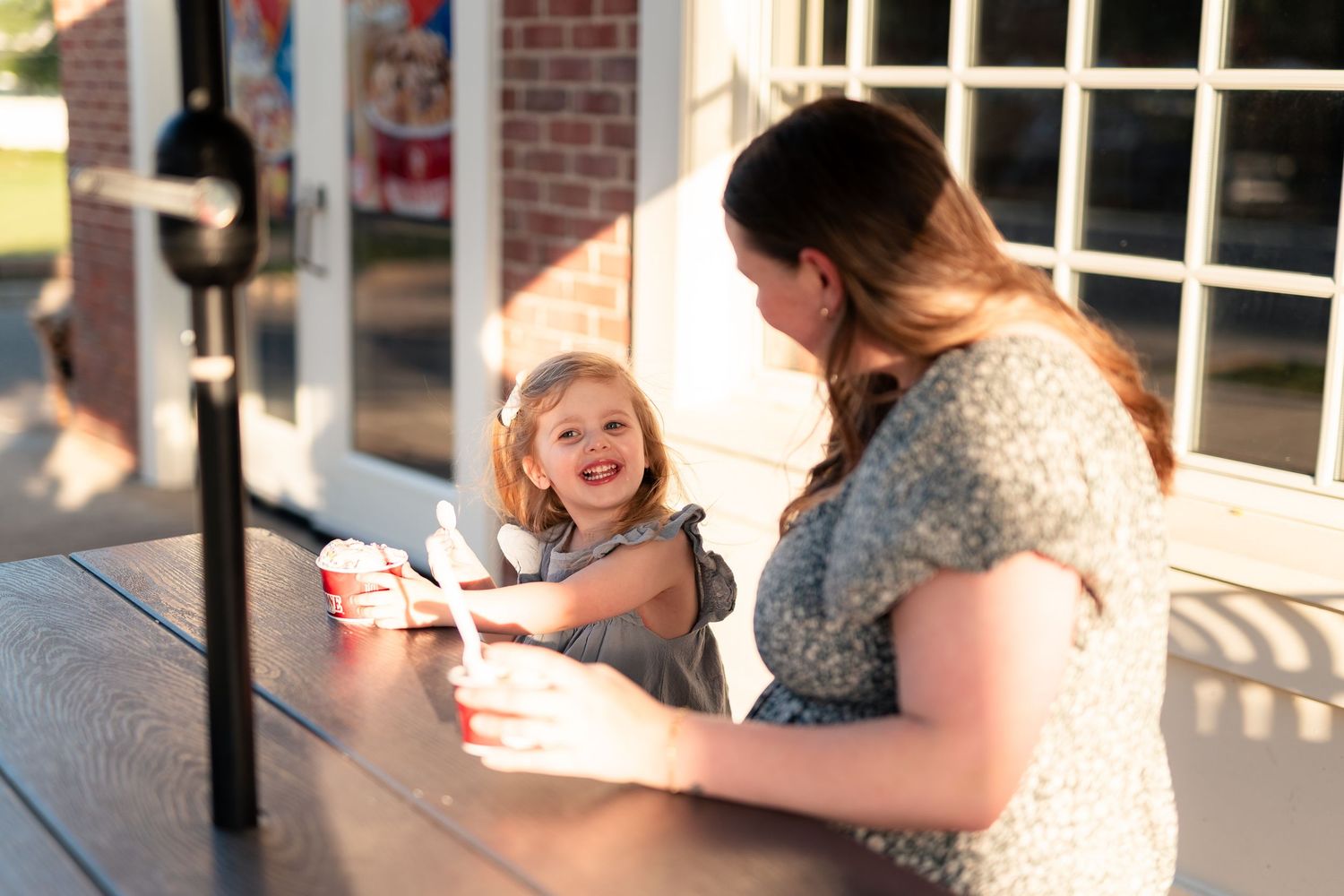 A young girl smiles while sharing ice cream with a woman at an outdoor table in warm golden light.