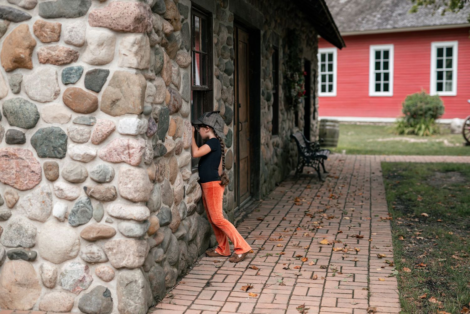 Woman in orange pants and black top leaning against a rustic stone building on a brick pathway.