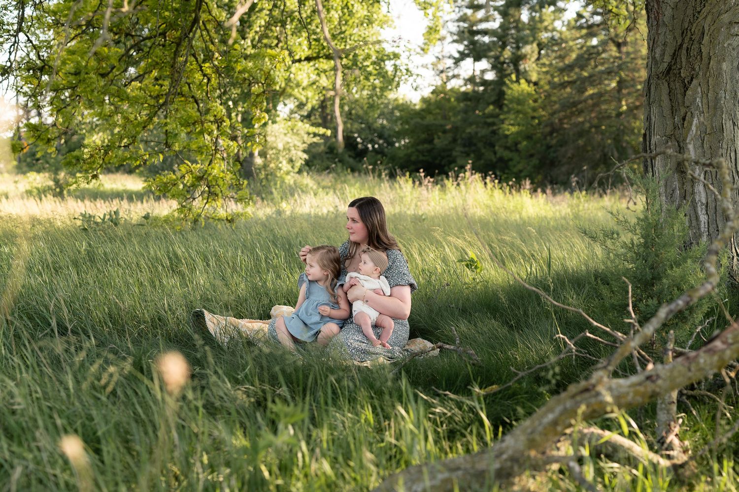 Mother sitting in lush green meadow under large tree with two young children on sunny day.