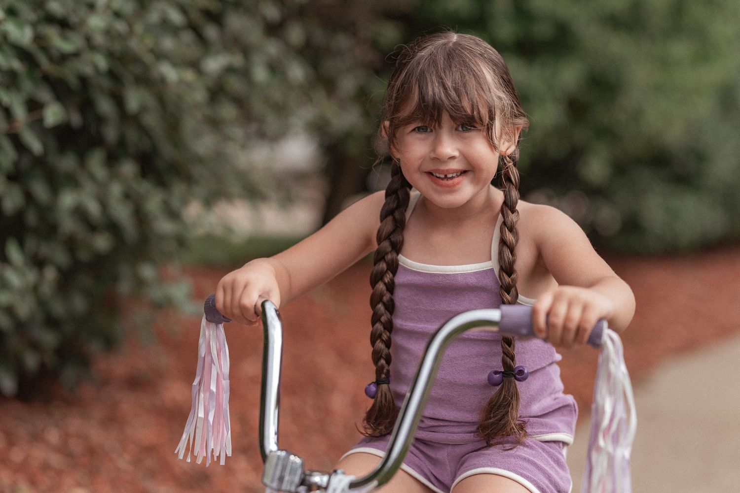 Child in purple tank top sitting on bike handlebars with white tassels outdoors in garden setting.