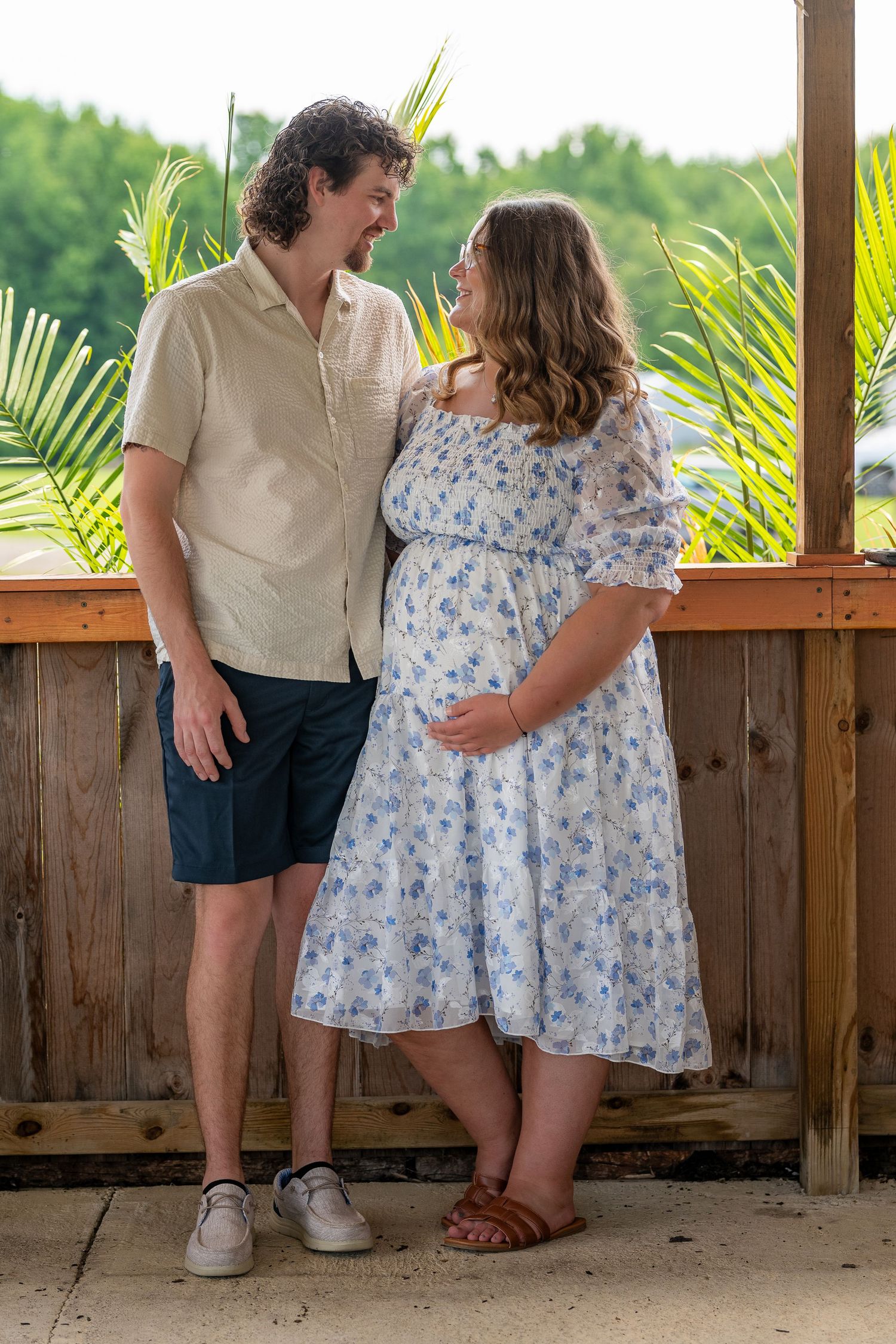 Pregnant couple smiling at each other on a wooden porch surrounded by tropical plants.