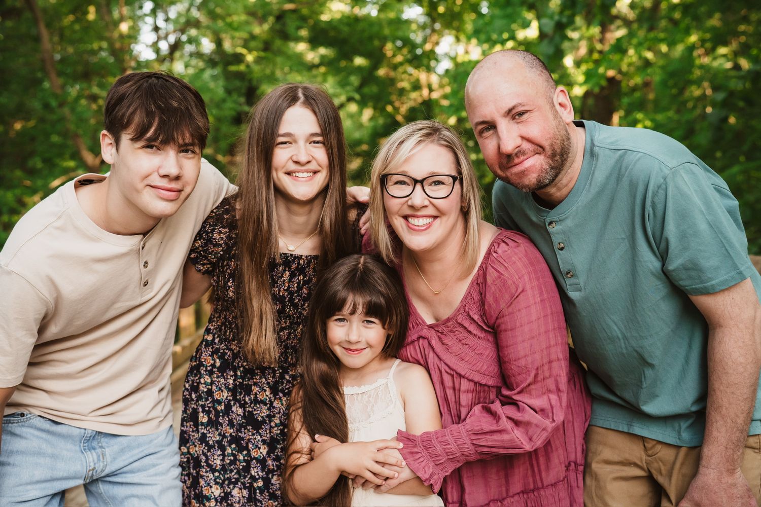 Happy family of five smiling together outdoors among lush green trees in a warm, candid portrait photo.