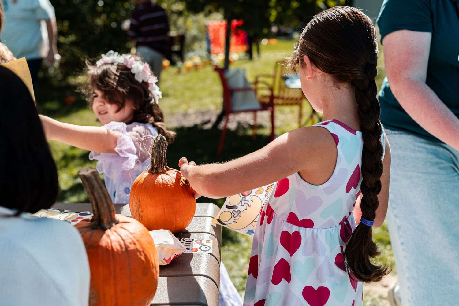 Two young girls reaching for orange pumpkins at an outdoor fall harvest festival event on a sunny day.