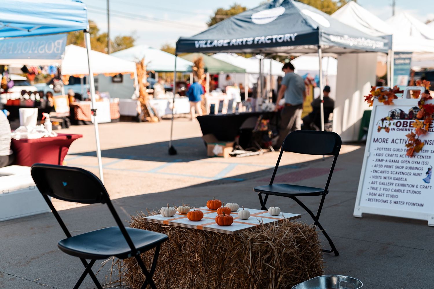 Outdoor fall market with hay bale display, pumpkins, and Village Arts Factory tent in background.