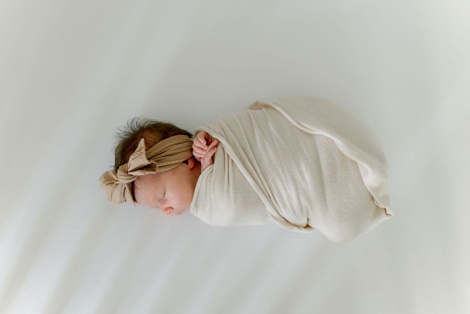 Swaddled newborn baby girl sleeps peacefully wearing a tan bow headband on white background.