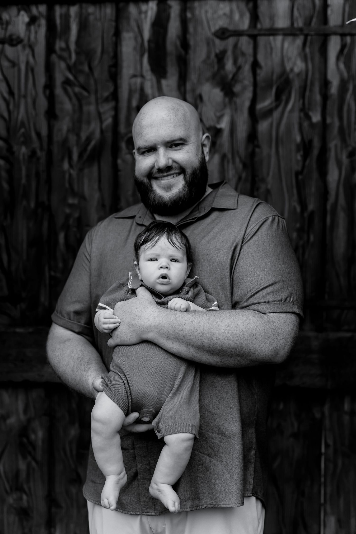 A smiling bald bearded man holds a newborn baby in a black and white portrait photo.