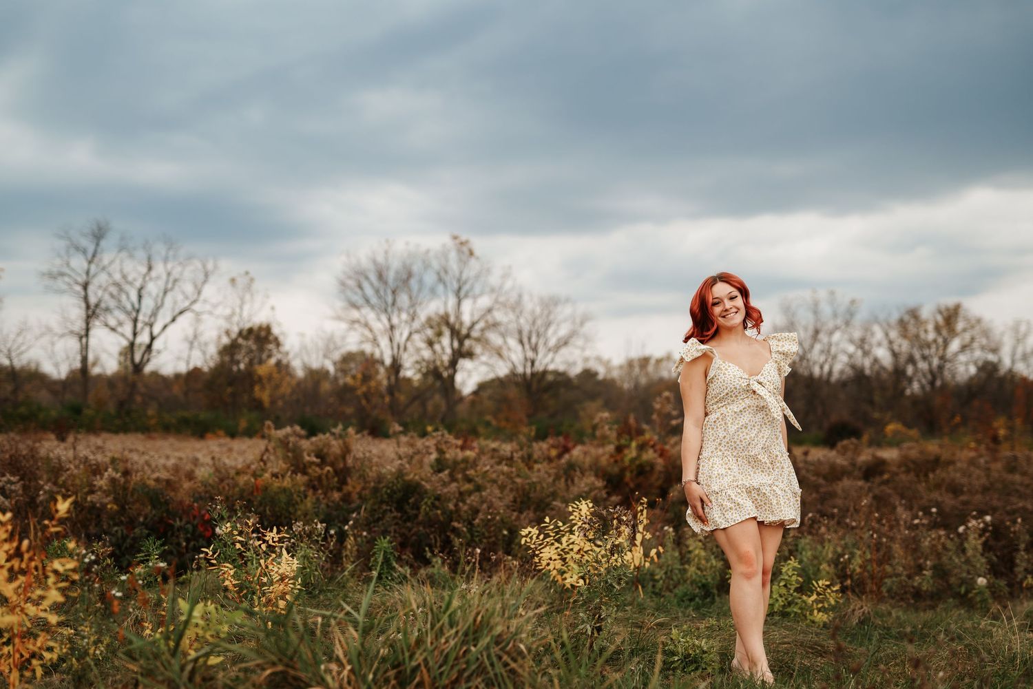 Red-haired woman in white dress standing in autumn field with bare trees and cloudy sky backdrop.