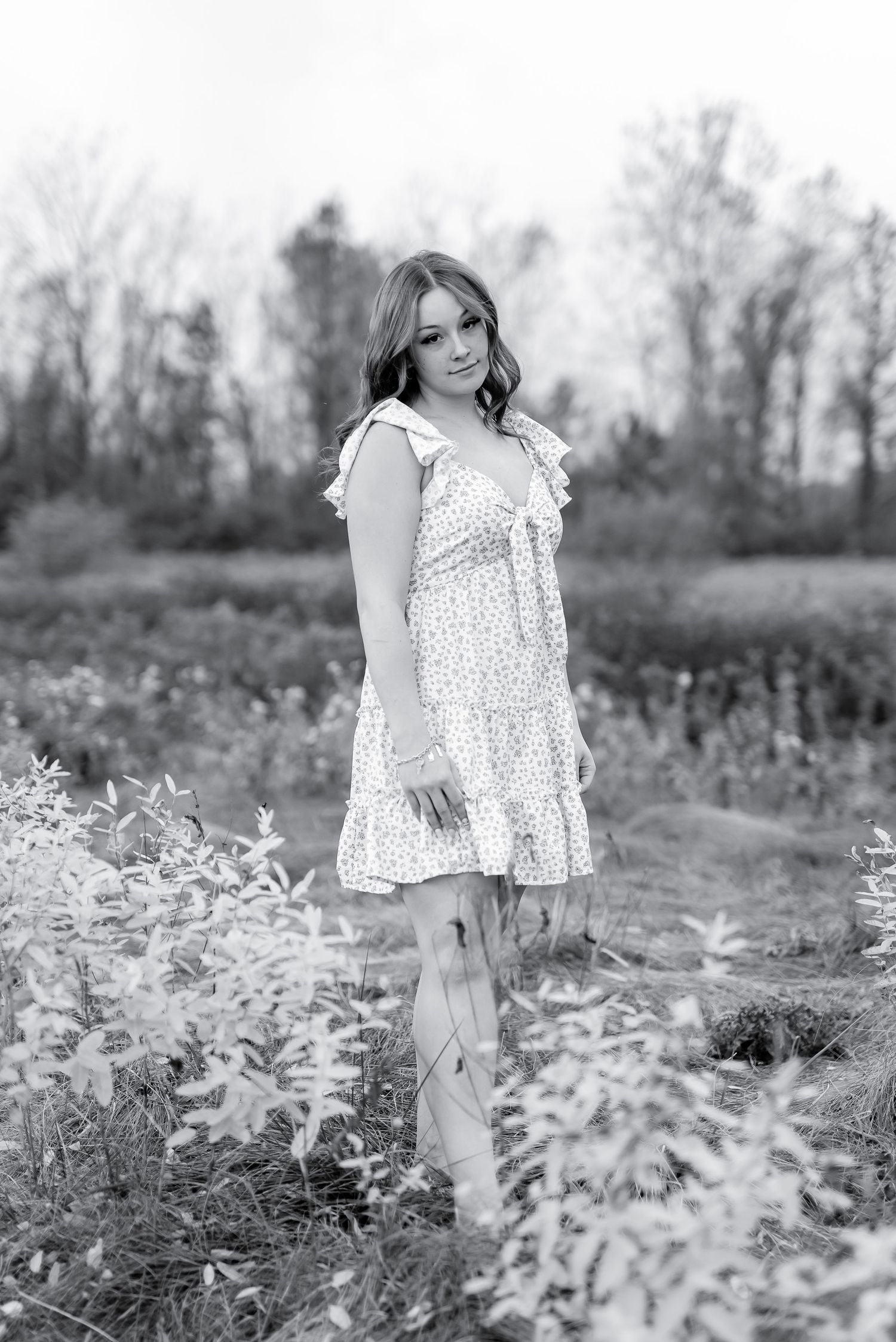 Black and white portrait of young woman in ruffle dress standing in outdoor field.