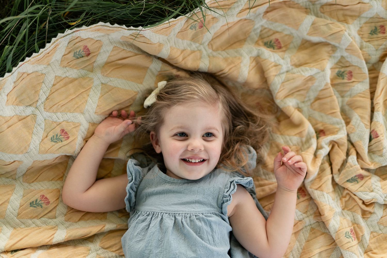 Smiling toddler girl in blue dress lying on yellow vintage quilt outdoors in grass, looking up happily.