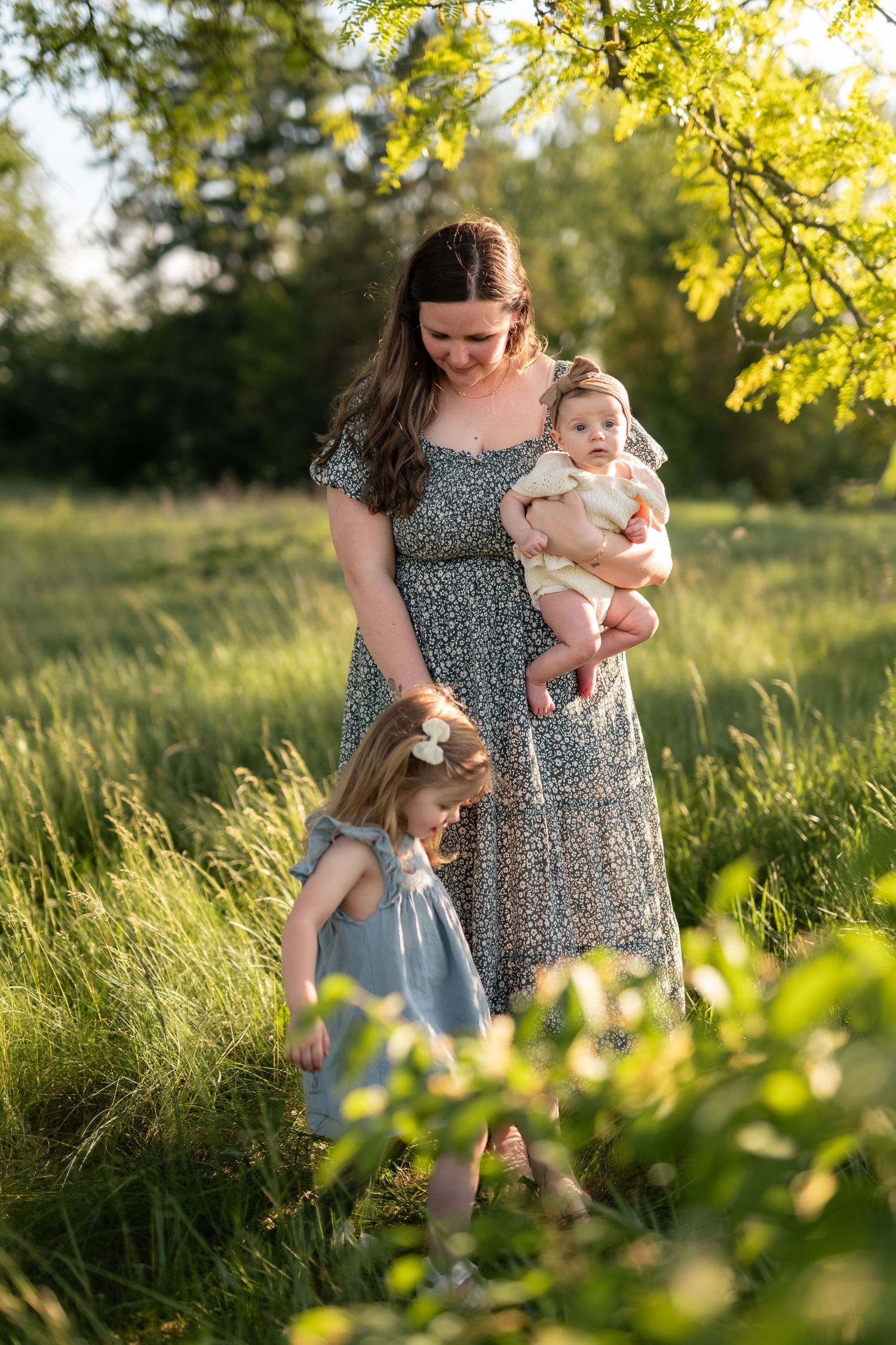 A mother in a floral dress holds her baby while her toddler daughter explores a sunlit green meadow.