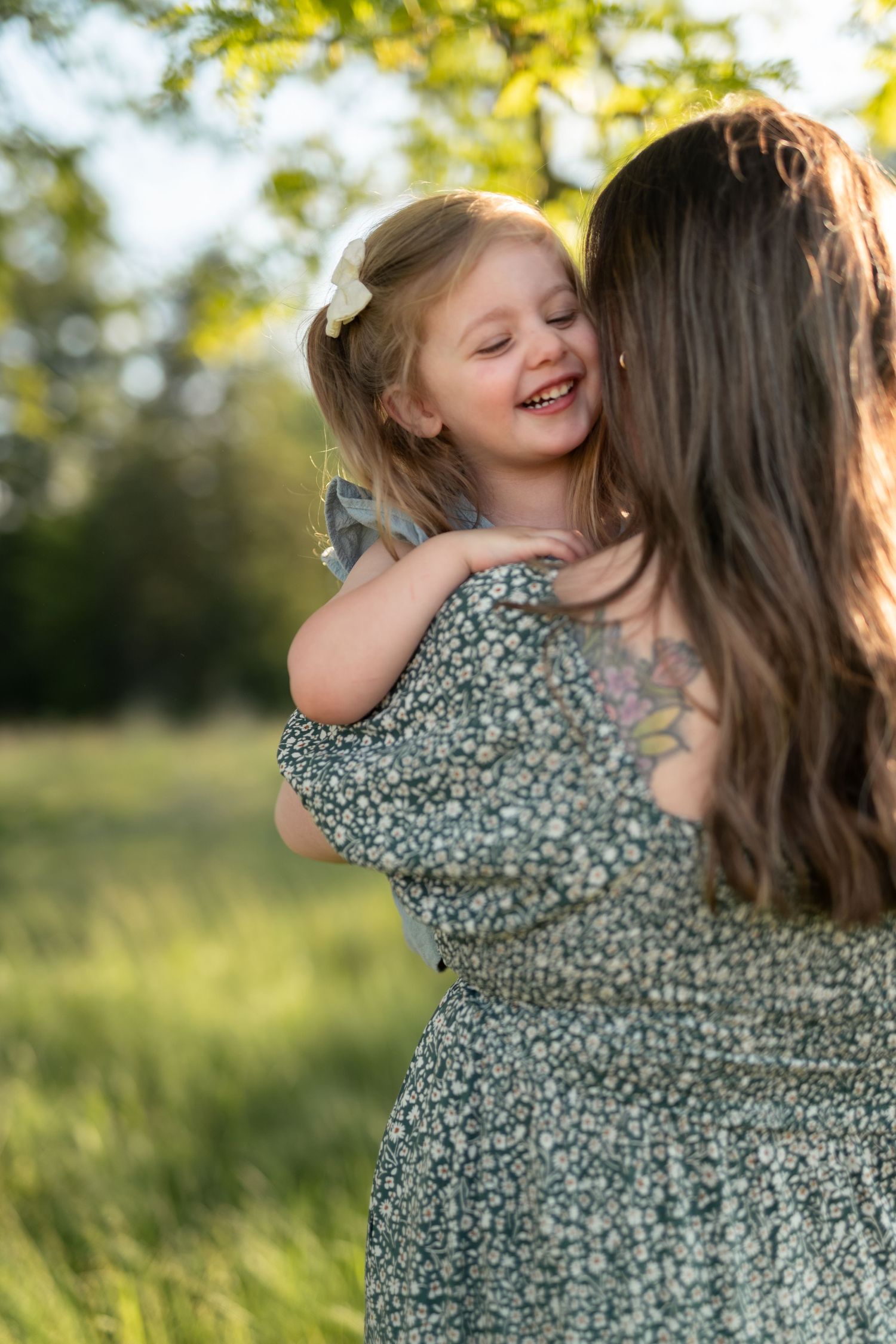 A mother in a floral dress holds her laughing toddler daughter outdoors in a sunlit green field.