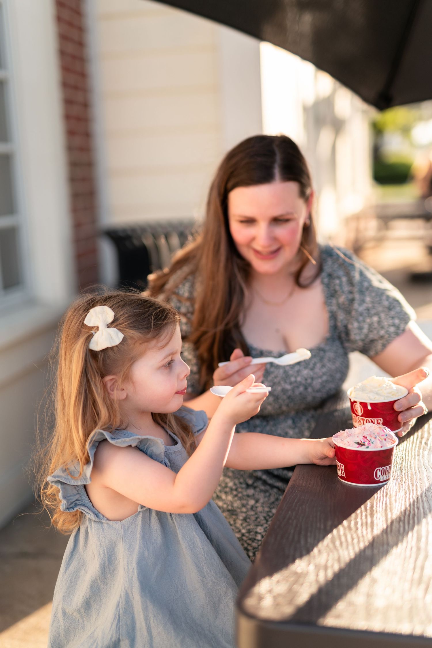 A mother and young daughter share ice cream together at an outdoor table on a sunny day.