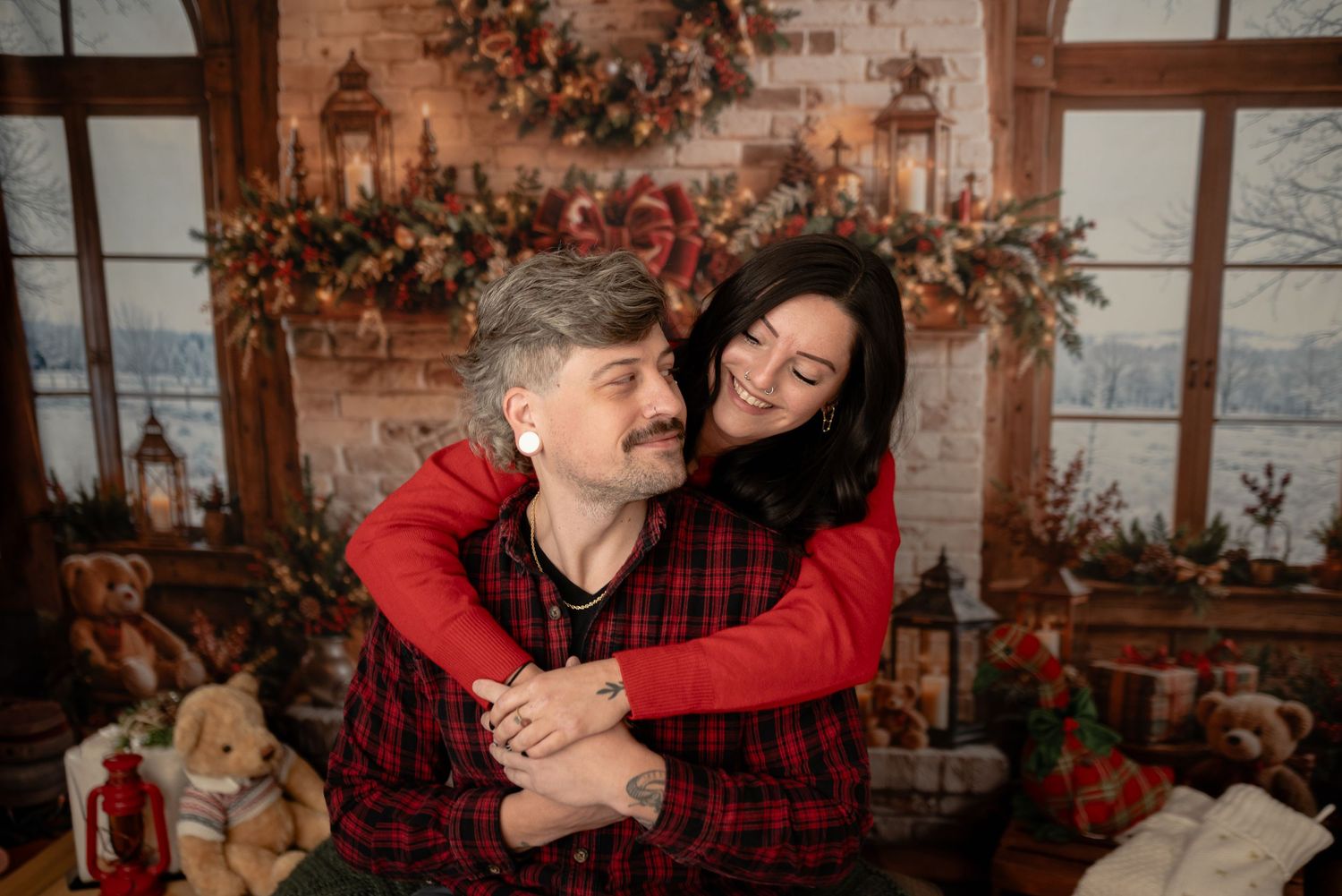 Smiling couple in red plaid and red sweater embrace in a cozy Christmas photo studio set with garlands, lanterns, and teddy bears.