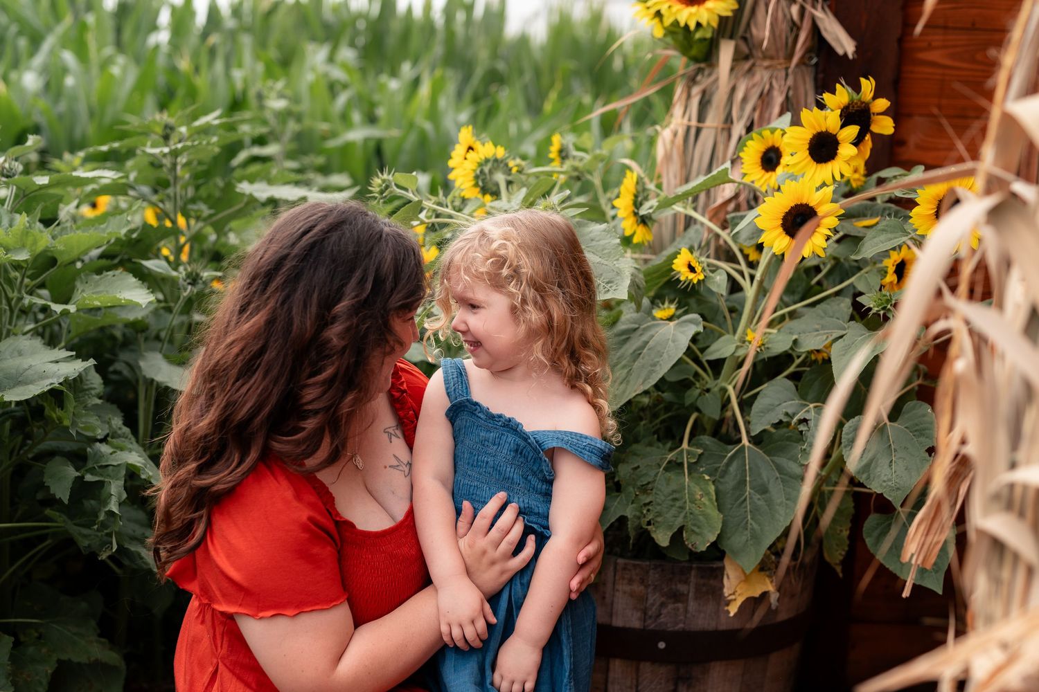 A mother in a red dress smiles at her toddler girl in a blue dress among sunflowers in a garden.
