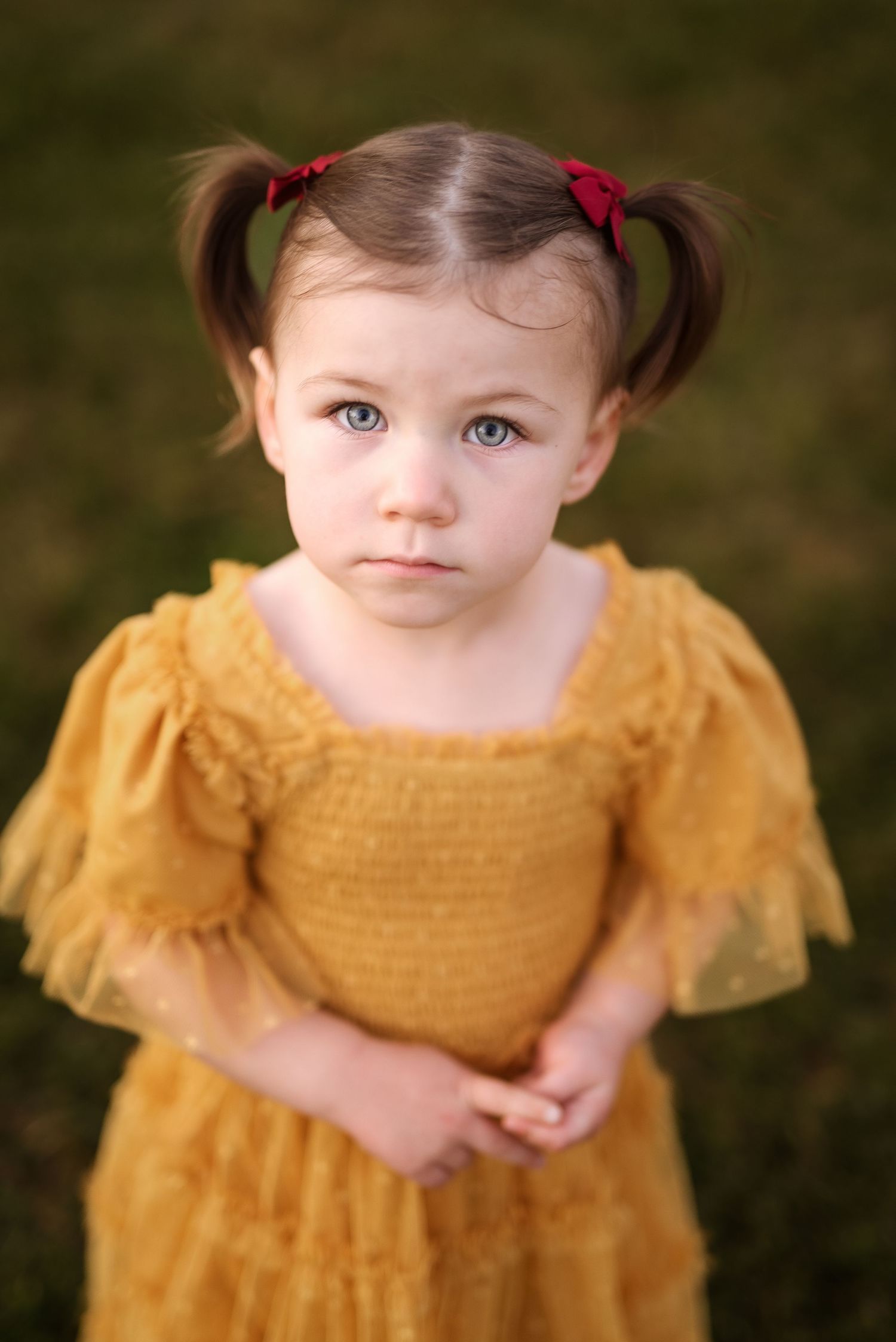 A young child in a mustard yellow dress with red hair bows poses against a dark background.