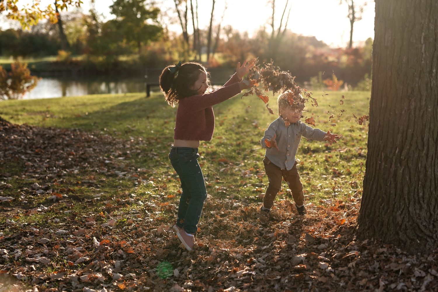 Two people playing and jumping in a pile of fallen autumn leaves during golden hour at a park.