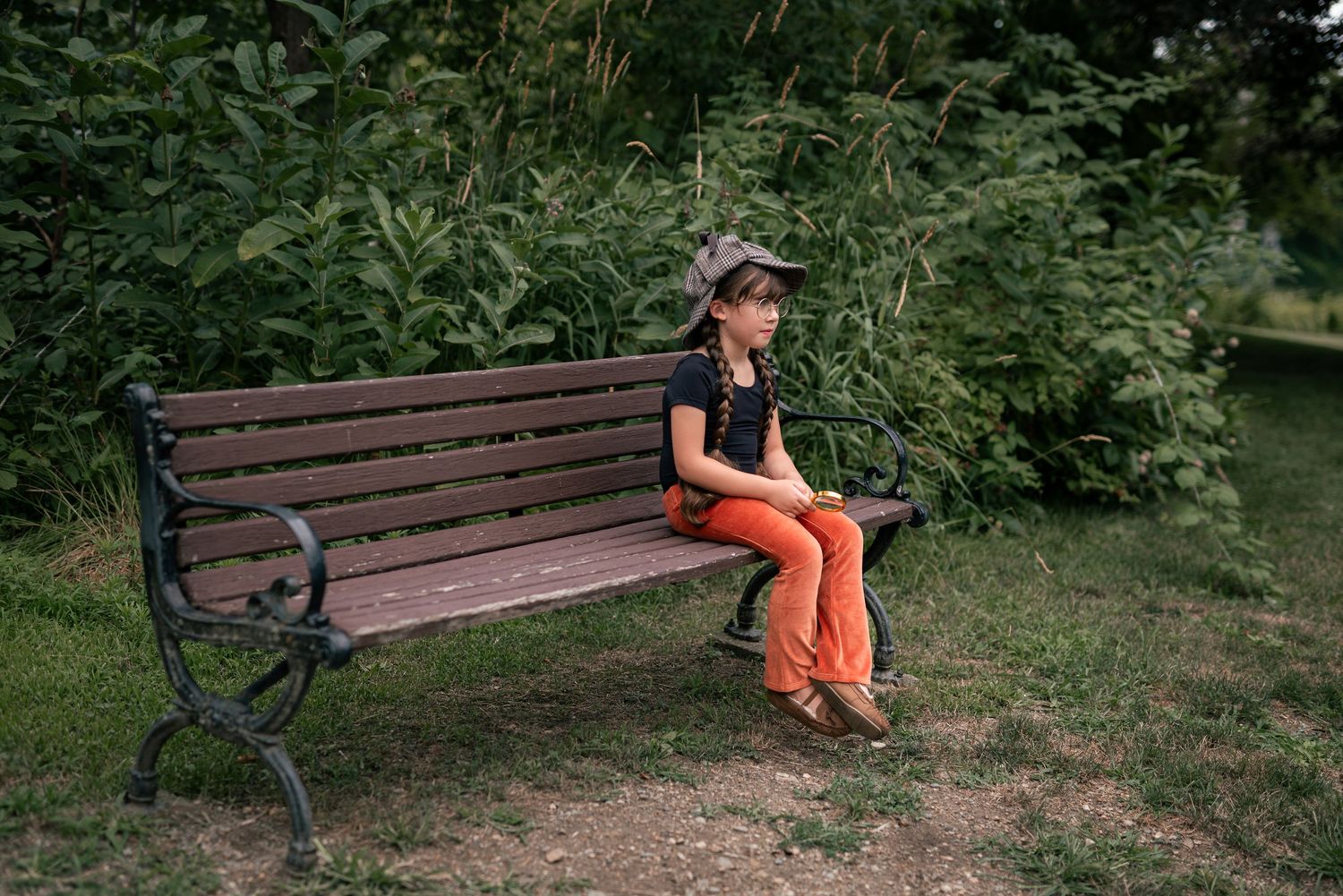 A young girl in orange pants and black top sits alone on a wooden park bench surrounded by lush green trees.