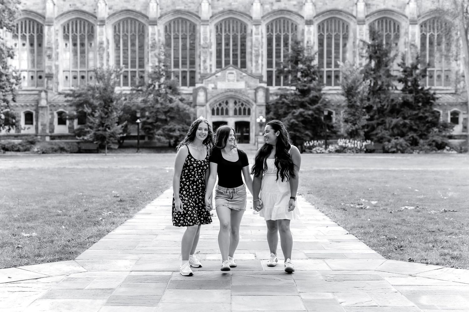 Students walk across a college campus pathway with Gothic-style architecture and green lawns in the background.
