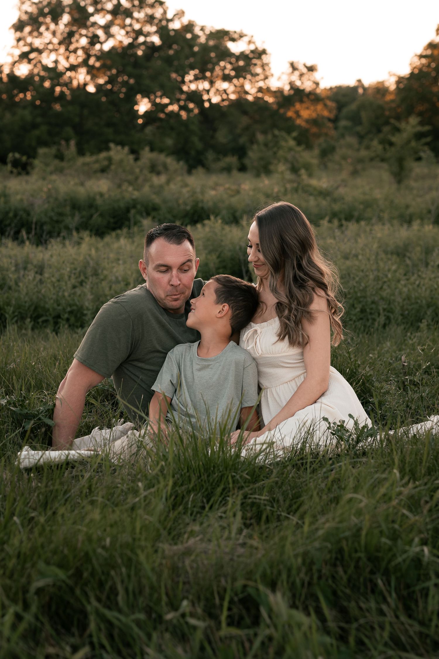 A family of three sits together in a lush green field at golden hour, sharing a tender moment.