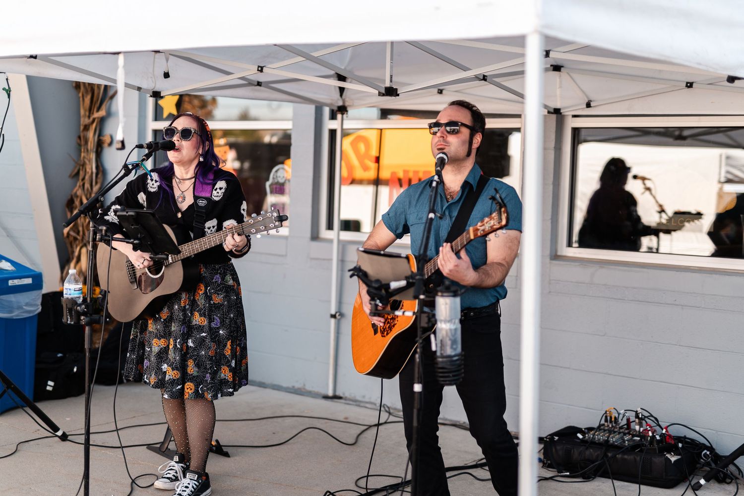 Male and female acoustic guitar duo performing live outdoor concert under white canopy tent at dusk.
