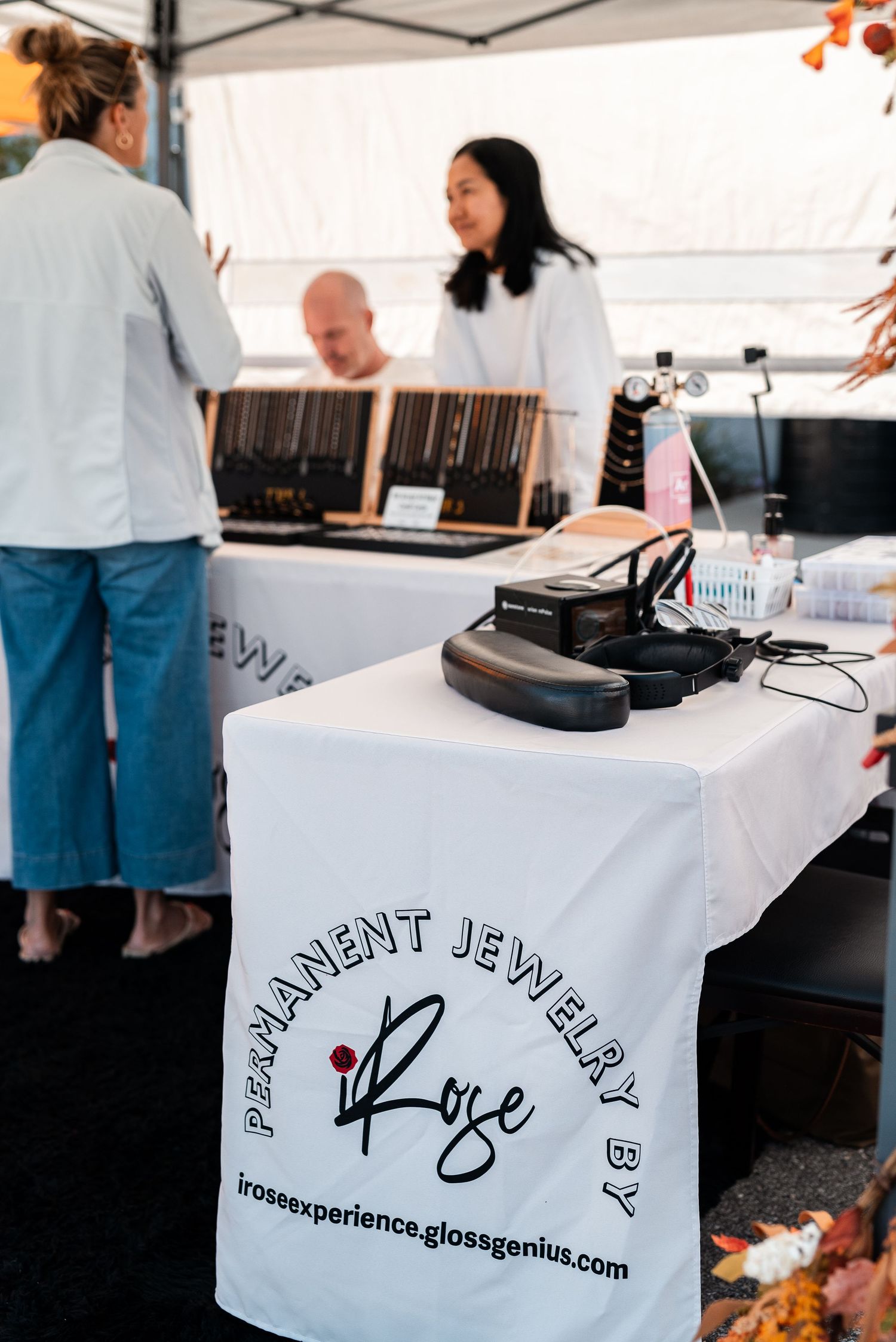 Permanent jewelry booth by iRose at an outdoor market with vendors and display cases visible.