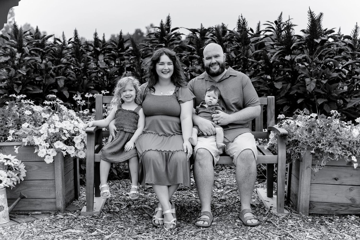 Family of four sitting on a bench surrounded by flower beds in a lush garden setting.